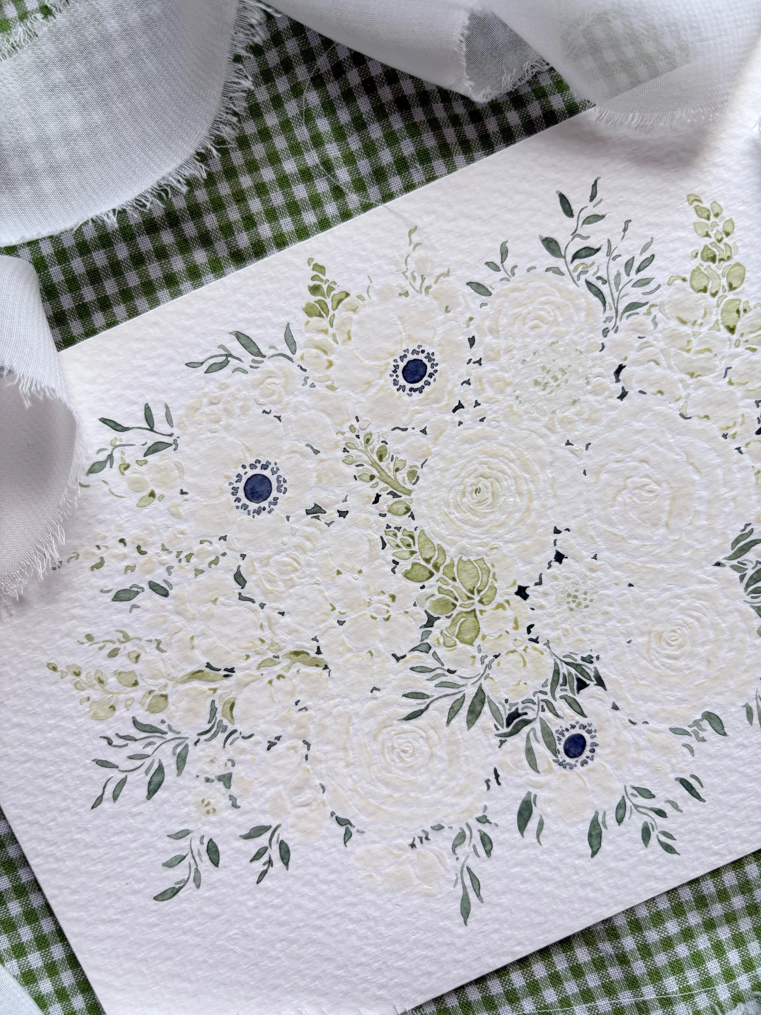 A decorative paper with a floral pattern featuring white and green roses and leaves, placed on a green and white checkered cloth, with a white cloth partially visible in the top left corner.