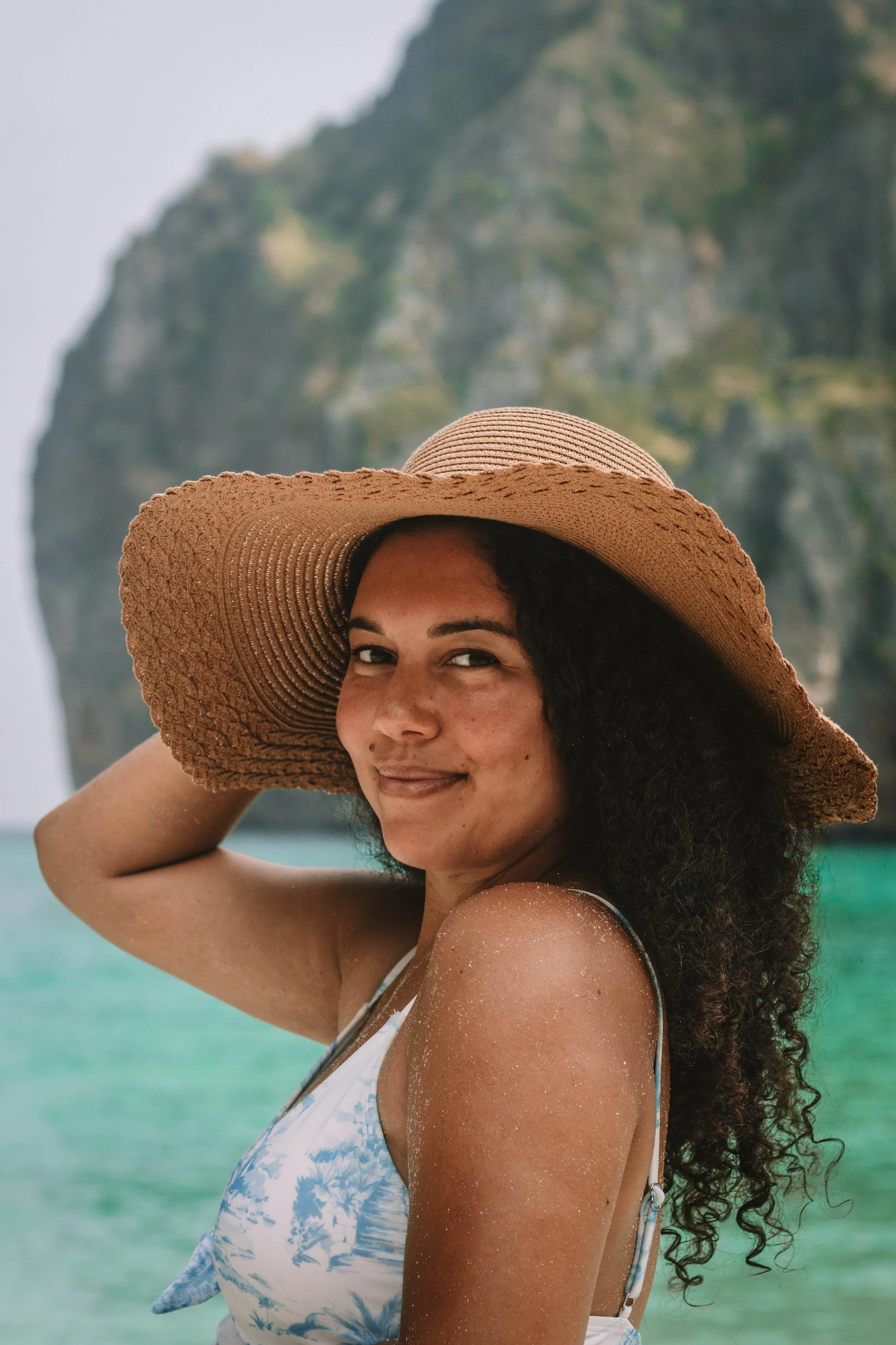 A travel UGC creator took this photo for her content creator portfolio. She has curly hair wearing a large straw hat and a blue and white swimsuit, smiling at the camera, standing on a beach with turquoise water and a green cliff in the background.