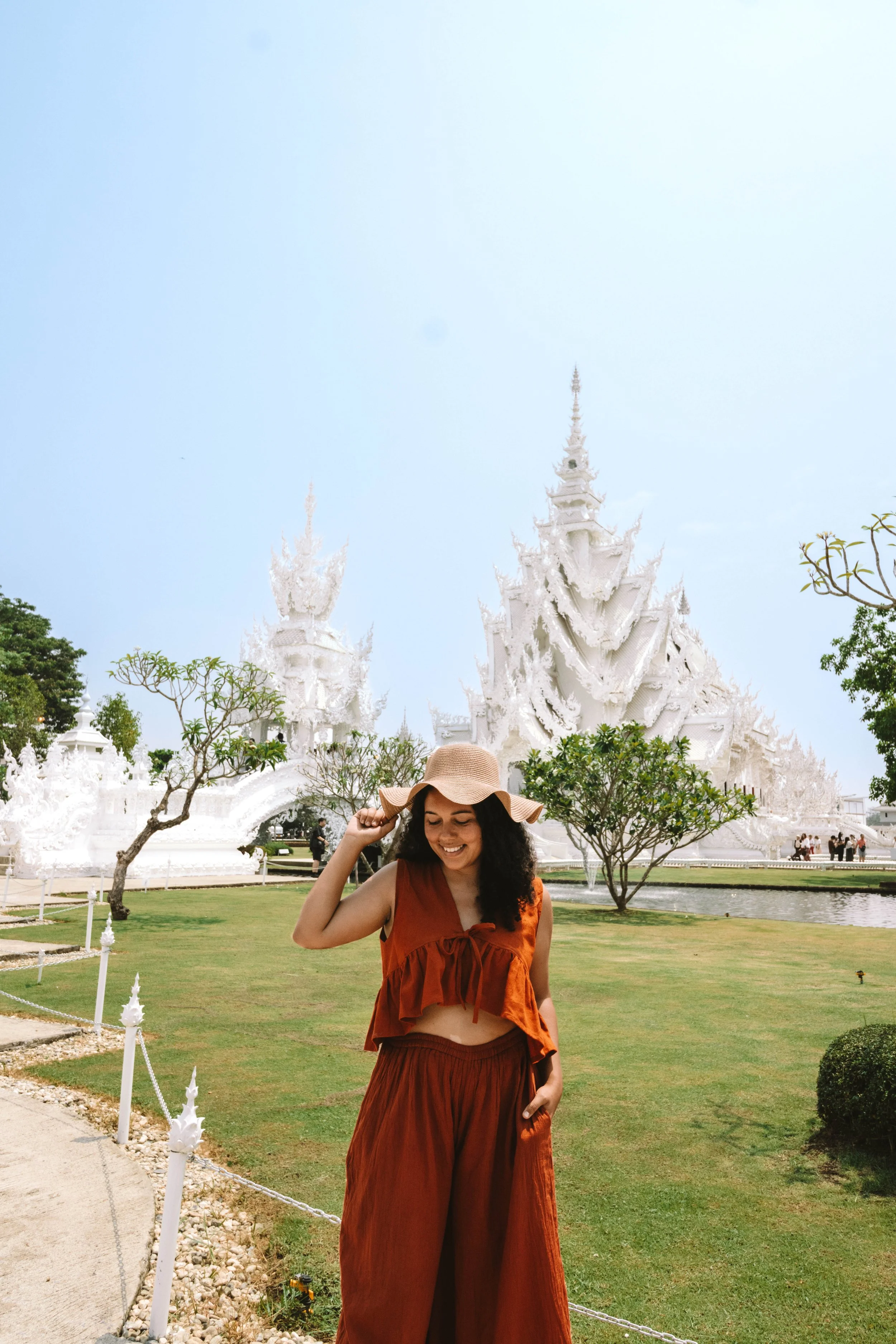 A woman who is traveling for work as a travel UGC creator wearing a wide-brimmed hat and an orange outfit standing in front of a white temple with spires during her travels to Thailand, surrounded by green trees and a lawn.