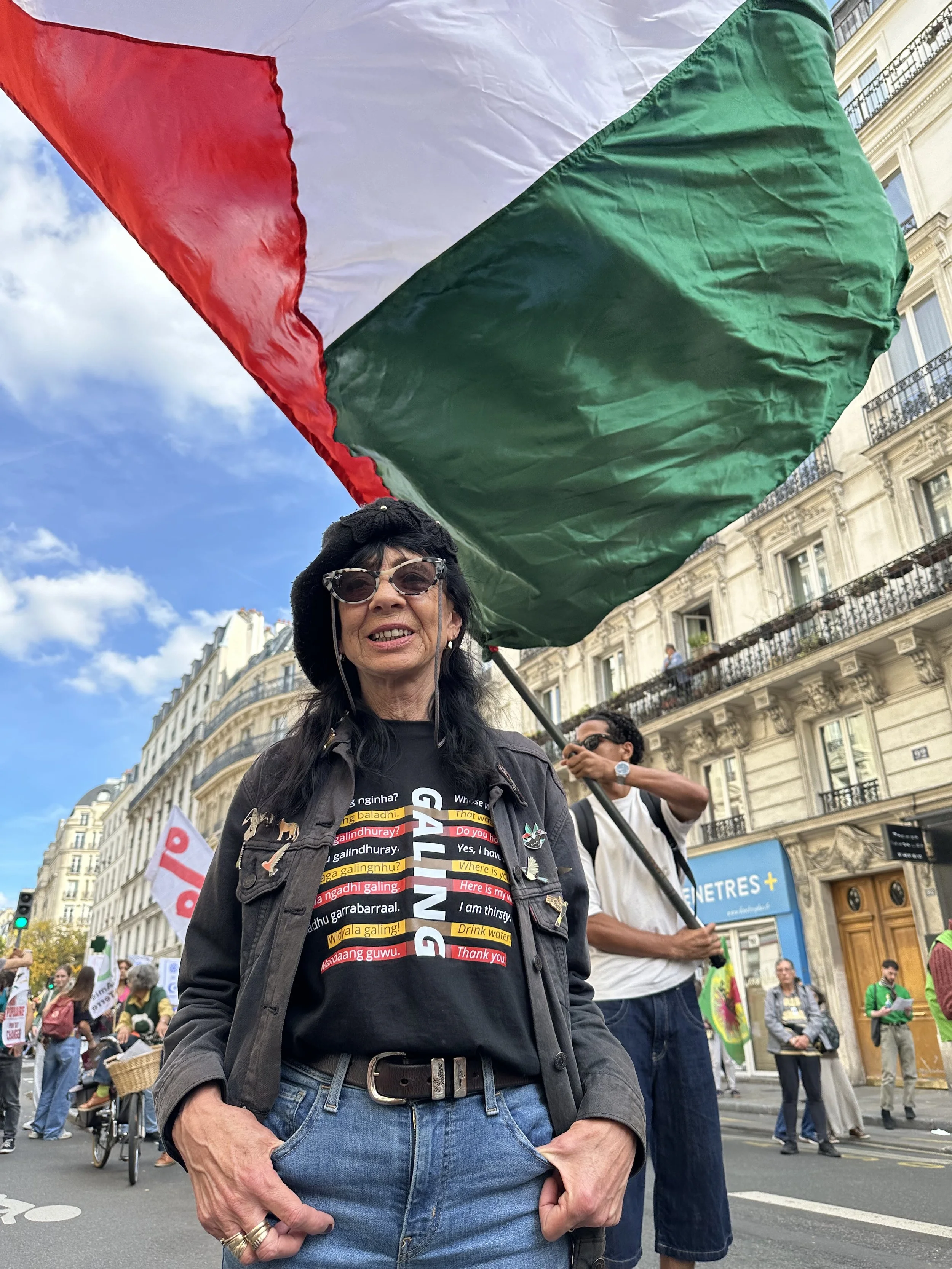 Image shows a thin, dark-haired woman standing. She wears T Shirt containing Wiradjuri word for water. In the background is a Palestinian flag, protest marchers and Parisian tenement housing.