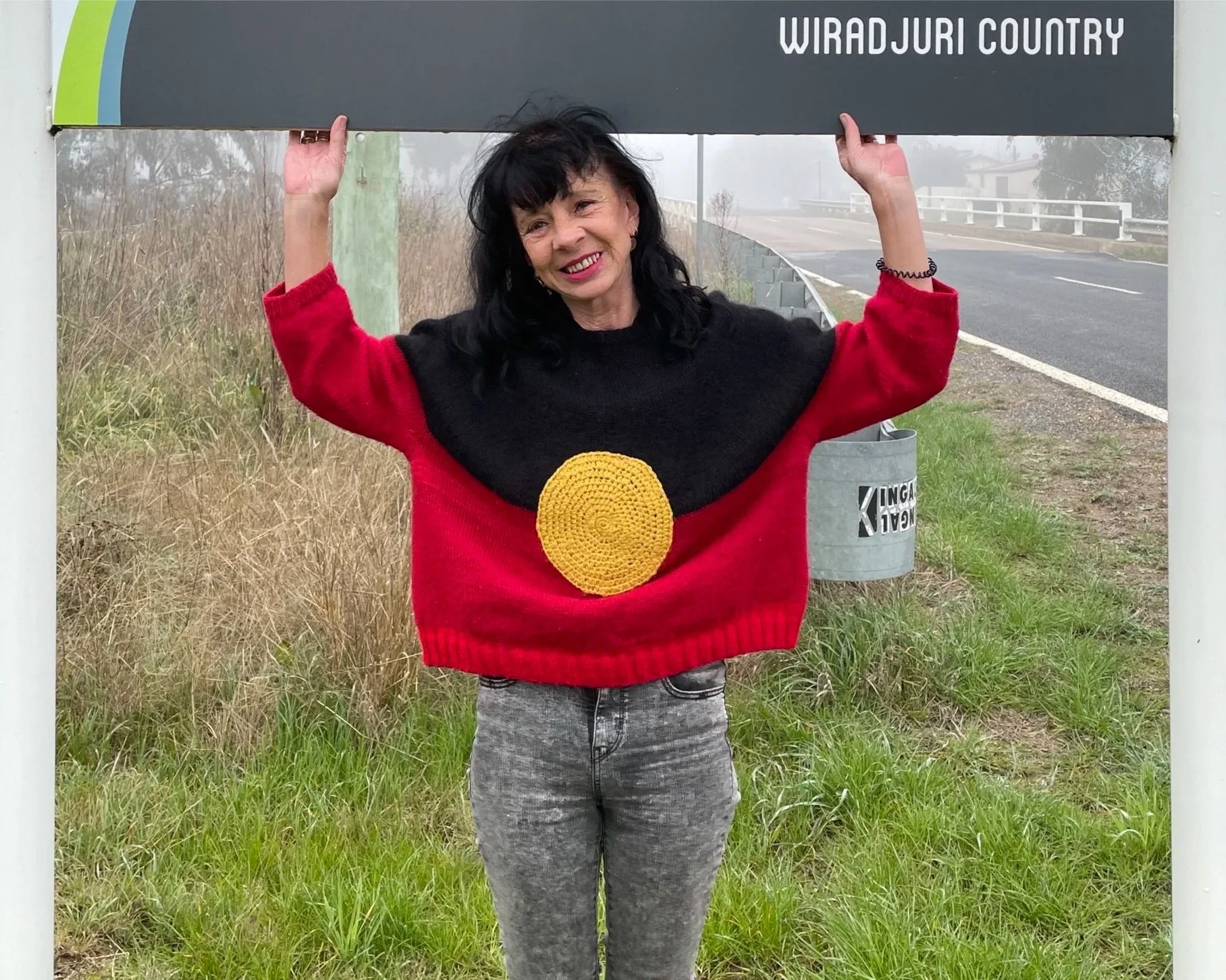 A slim woman with black hair, stands under a road sign reading  "Wiradjuri Country". She wears a wooly jumper in the colours of the Aboriginal flag and dark jeans. Her arms are raised as if to hold the sign. In the background is a small bridge.