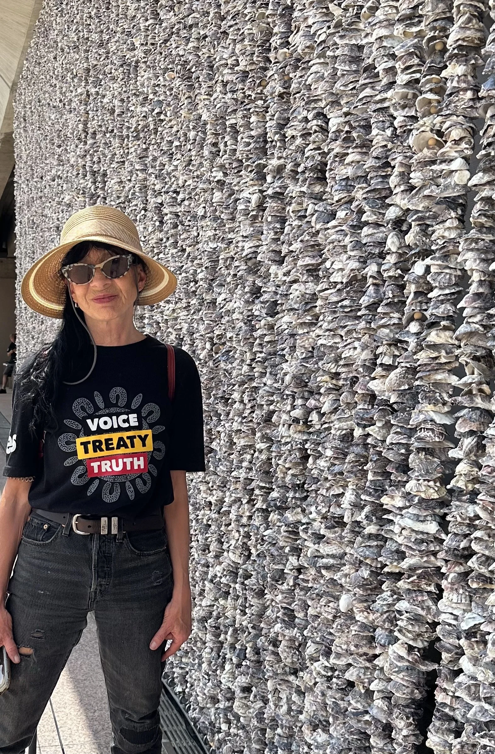 Image shows a thin, dark-haired woman looking at the camera. She is wearing a T Shirt with a logo that says Voice, Treaty, Truth.  The background is an art installation made up of vertically-hanging sea shells.