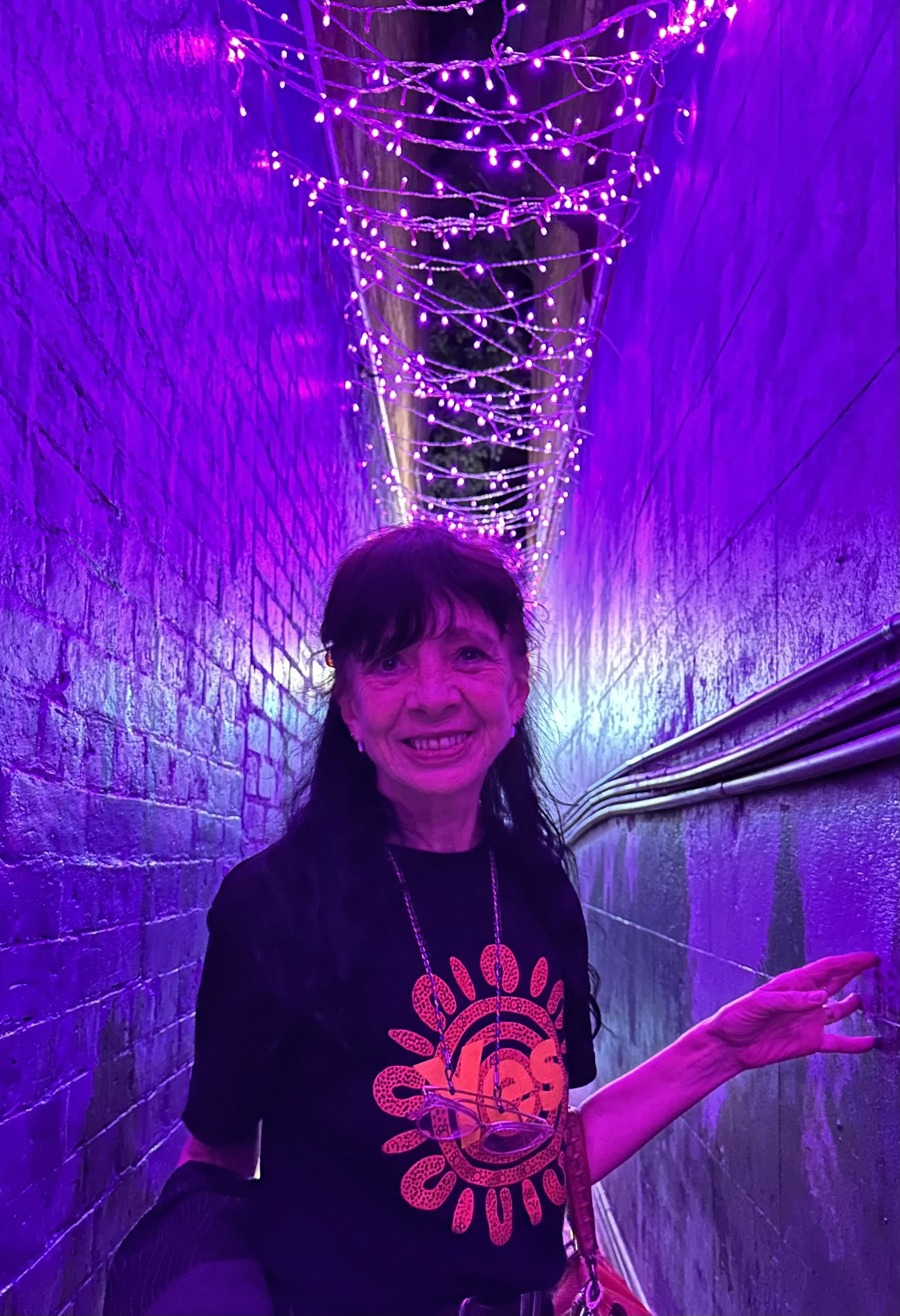 Image shows a thin, dark-haired woman smiling at the camera. She wears a black tshirt emblazoned with the word Yes. She stands between two brick walls forming a narrow corridor. It is night time and the area is bathed with soft purple light.