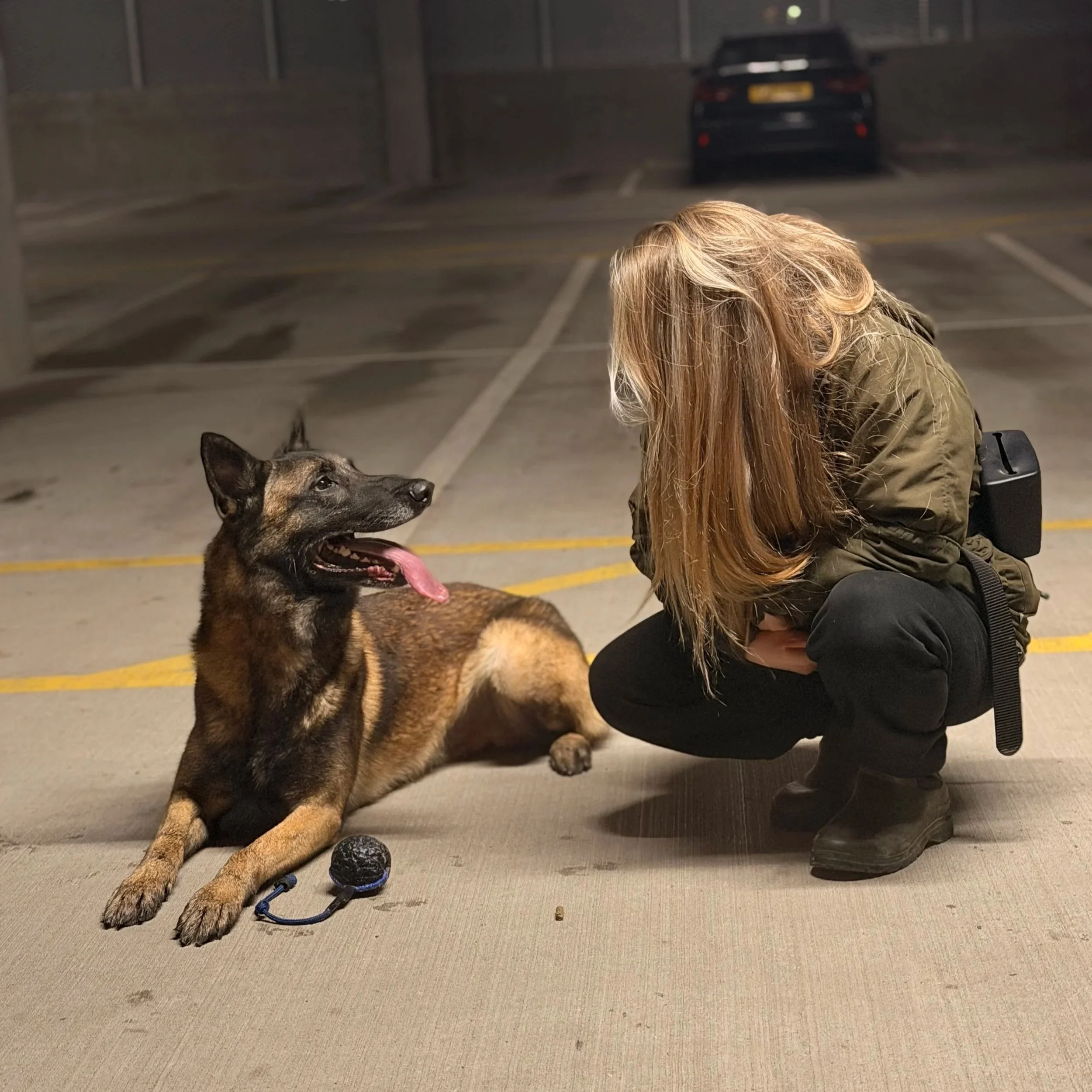 A woman crouching next to a seated Belgian Malinois dog in a parking garage at night.