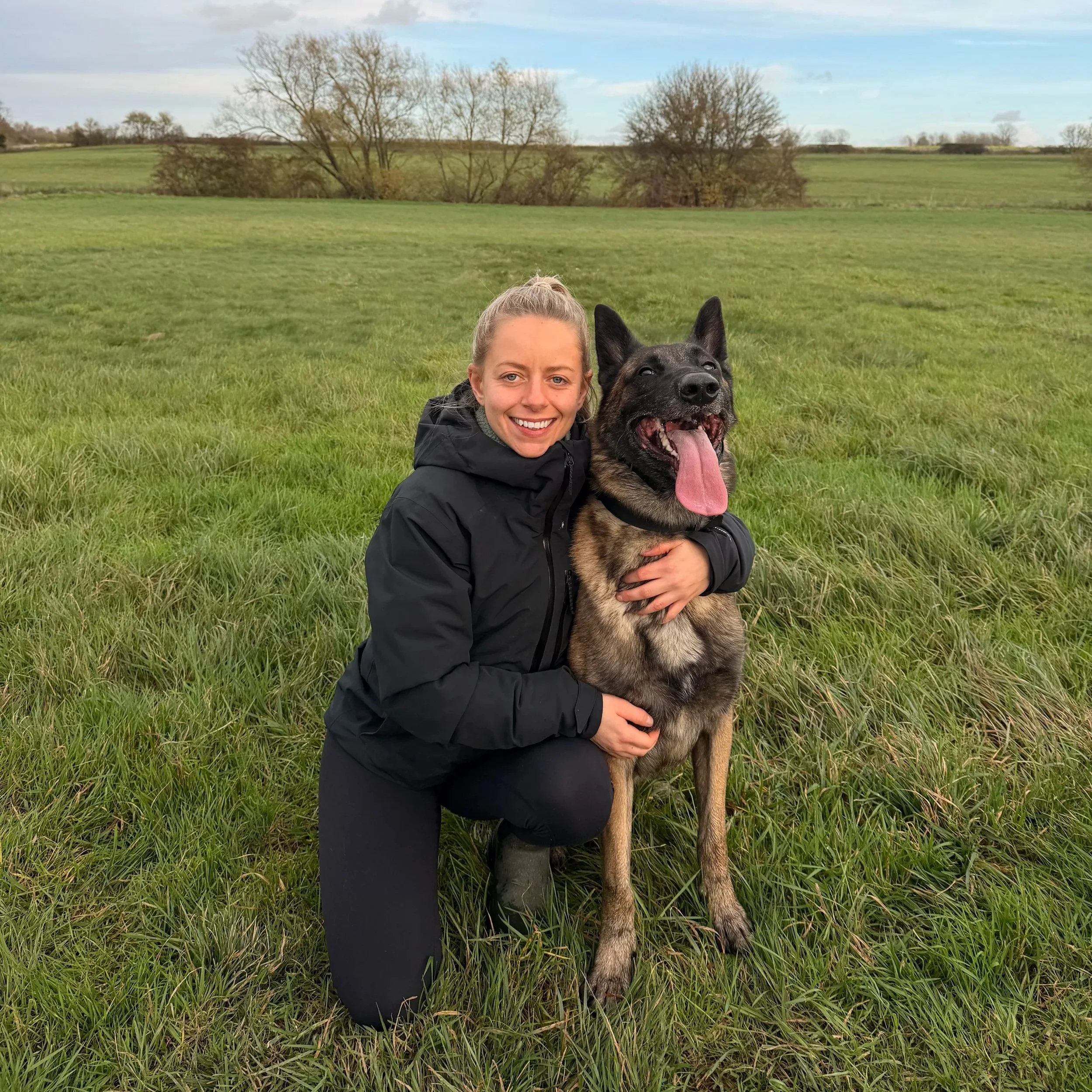 A woman kneeling on grass in an open field embracing a large German Shepherd dog with its tongue out. The sky is partly cloudy with trees in the background.