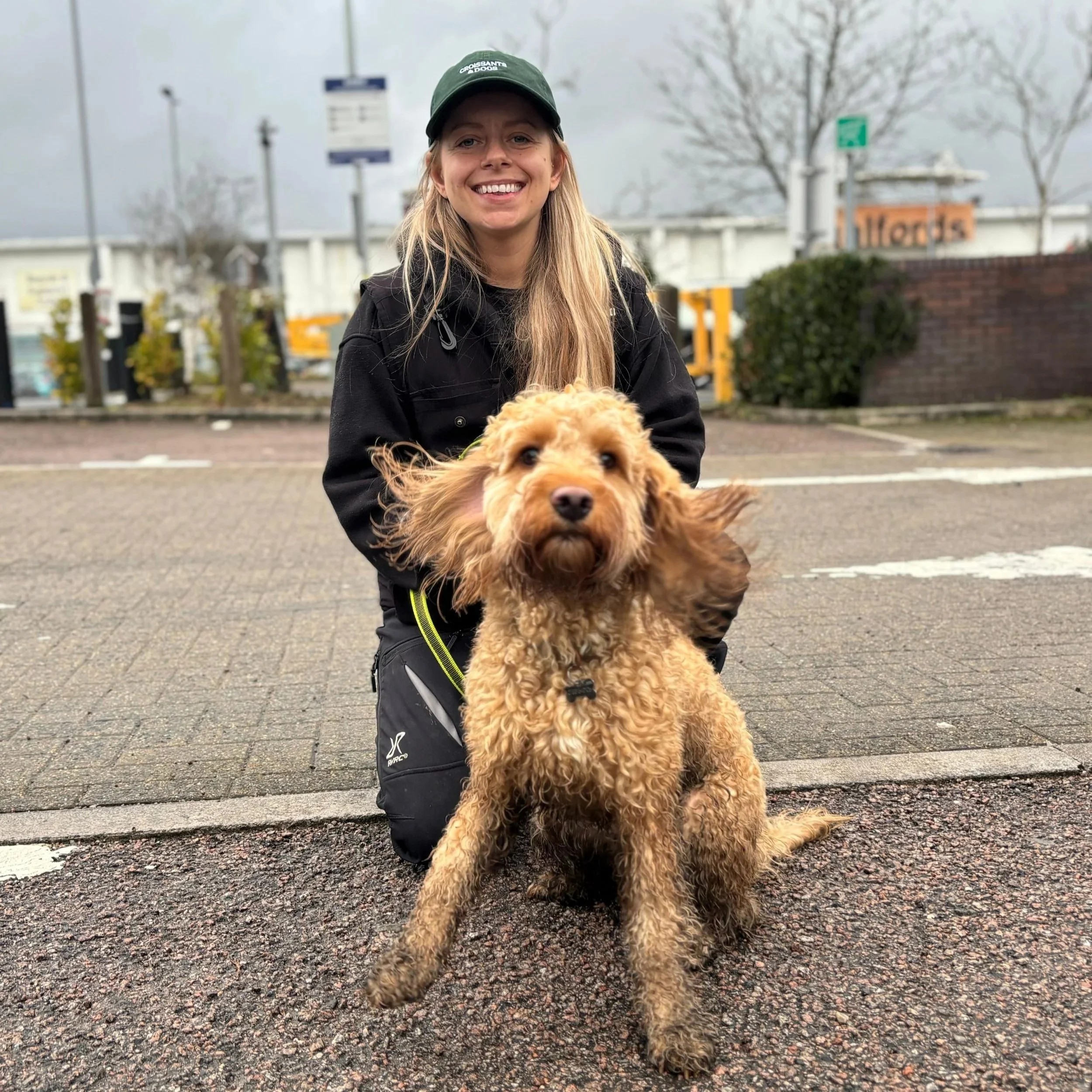 A woman in a green cap and black jacket kneeling on a sidewalk, smiling, with a curly-haired brown dog in front of her, outdoors on a cloudy day.