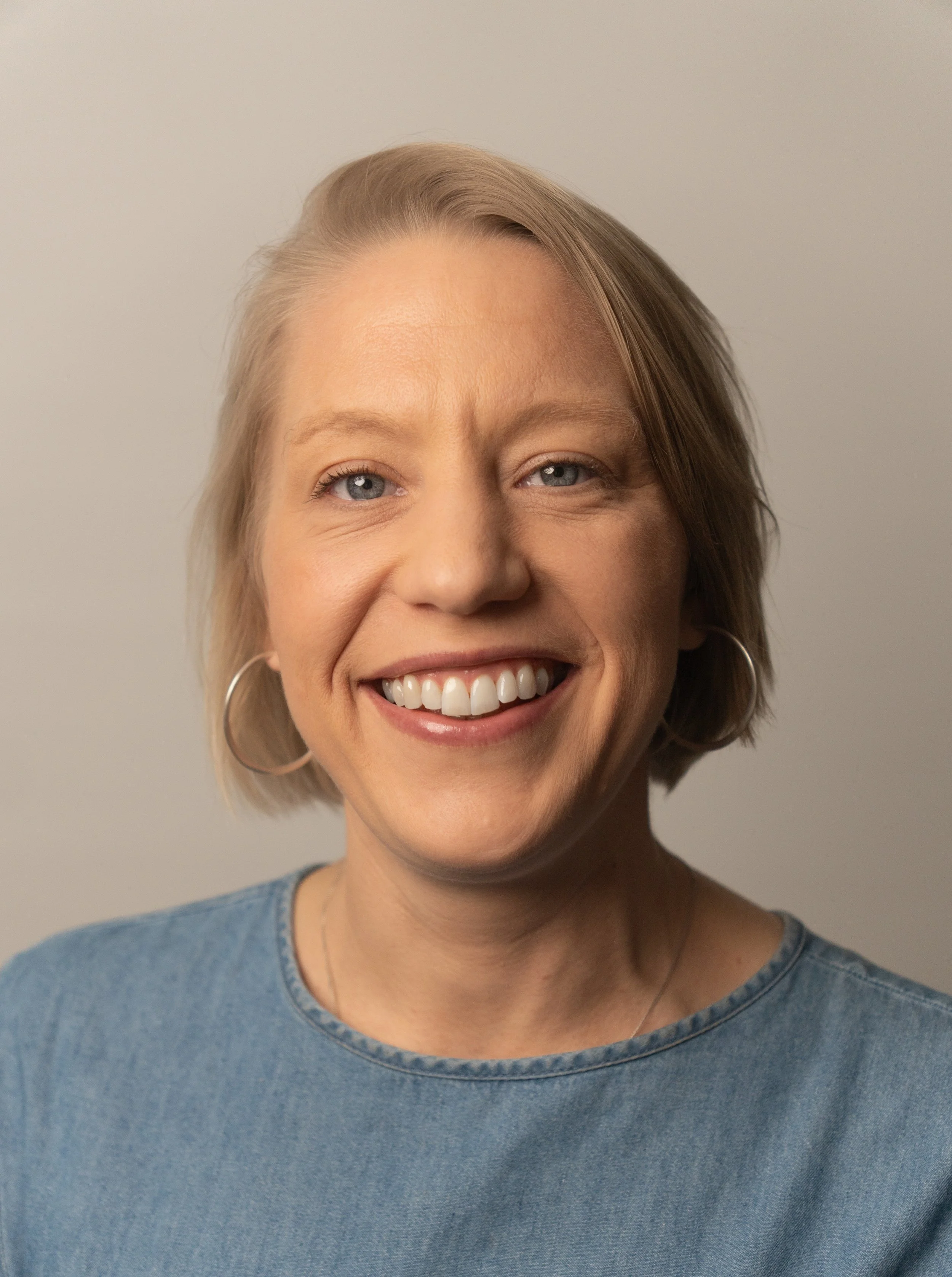 A woman with blonde hair smiling against a neutral wall background.