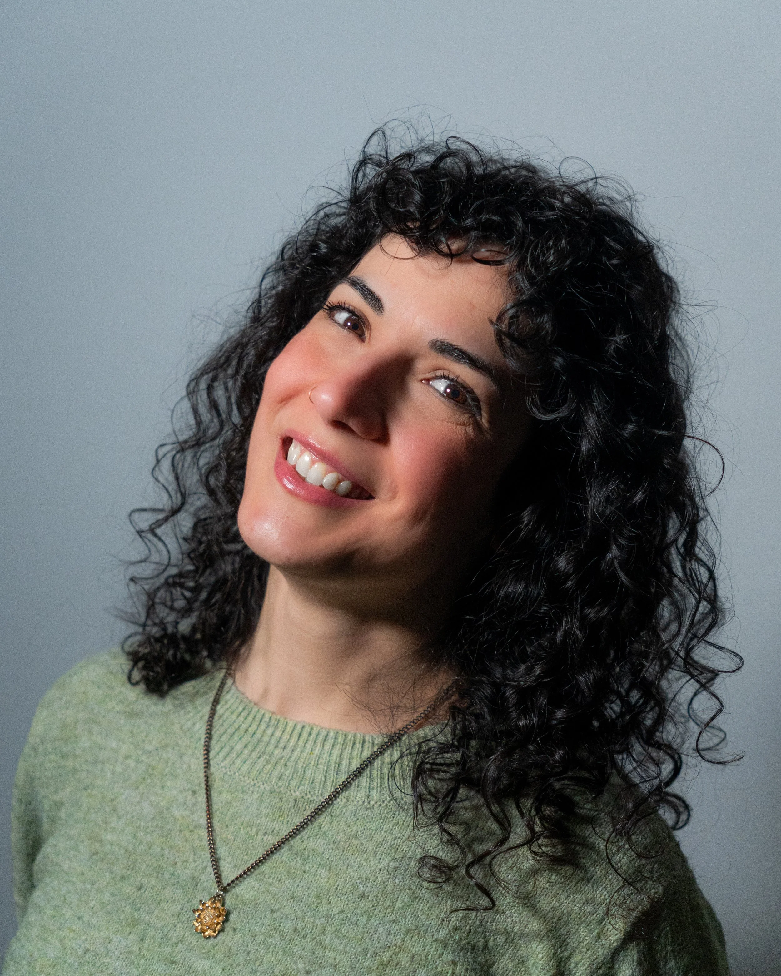 Portrait of a woman with curly black hair, smiling, wearing a turquoise top and a silver necklace with a large pendant, standing in front of a light-colored textured wall.