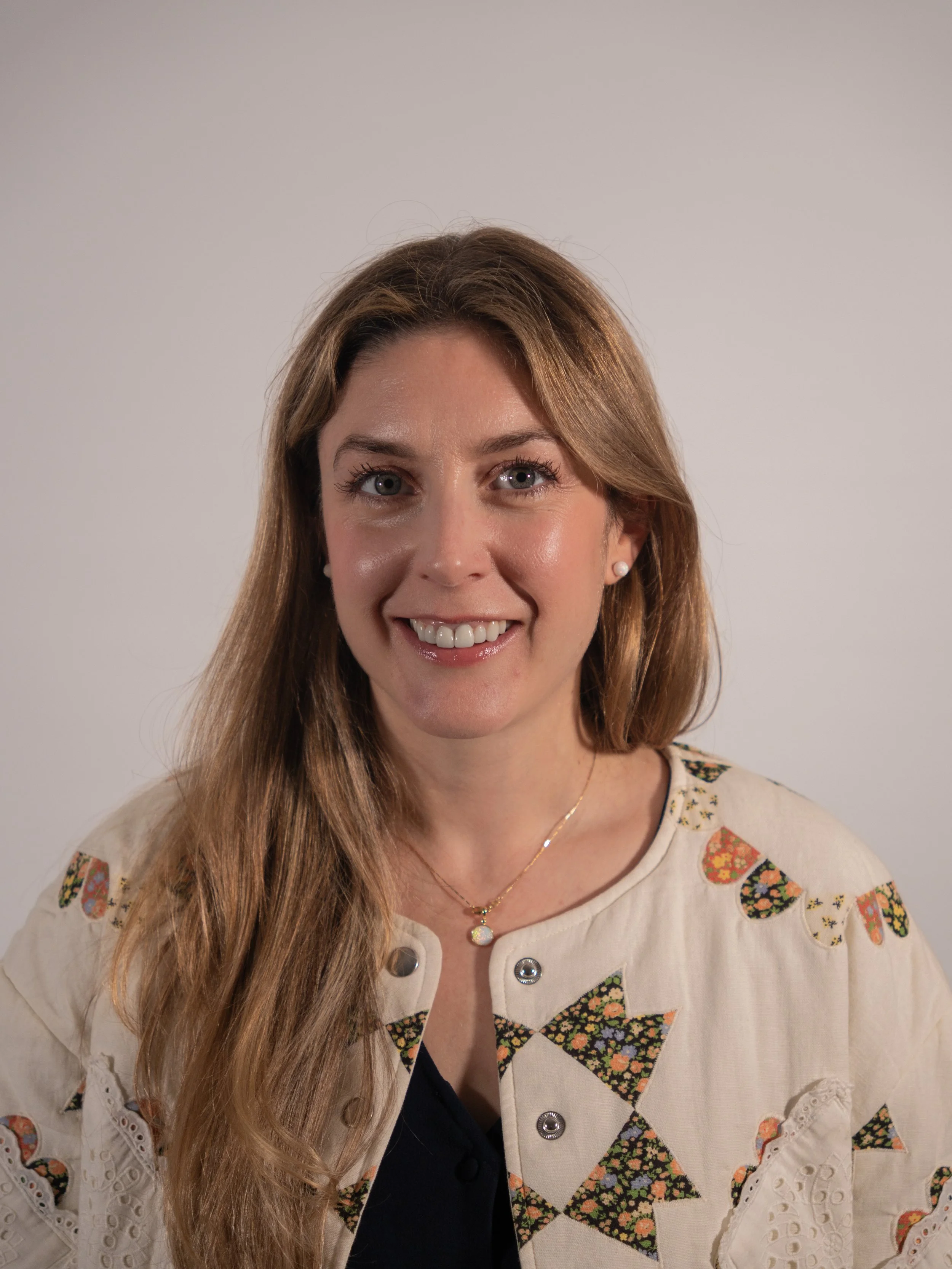 A young woman smiling with long, wavy hair, wearing a colorful sweater with pink and green patterns, standing against a textured neutral background.