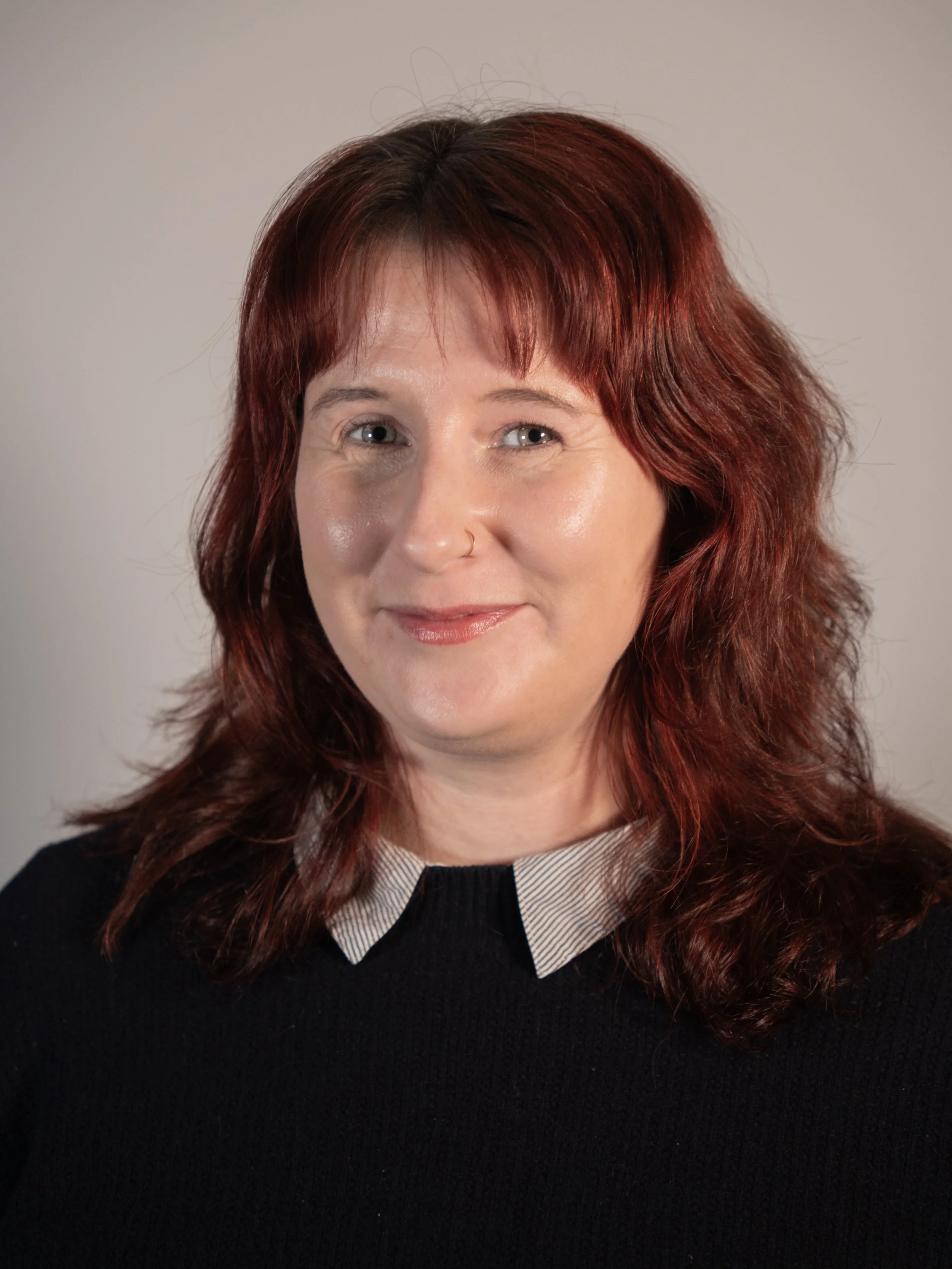 A woman with shoulder-length dark red hair, smiling, wearing a yellow button-up shirt with a black top underneath, standing against a beige textured wall.