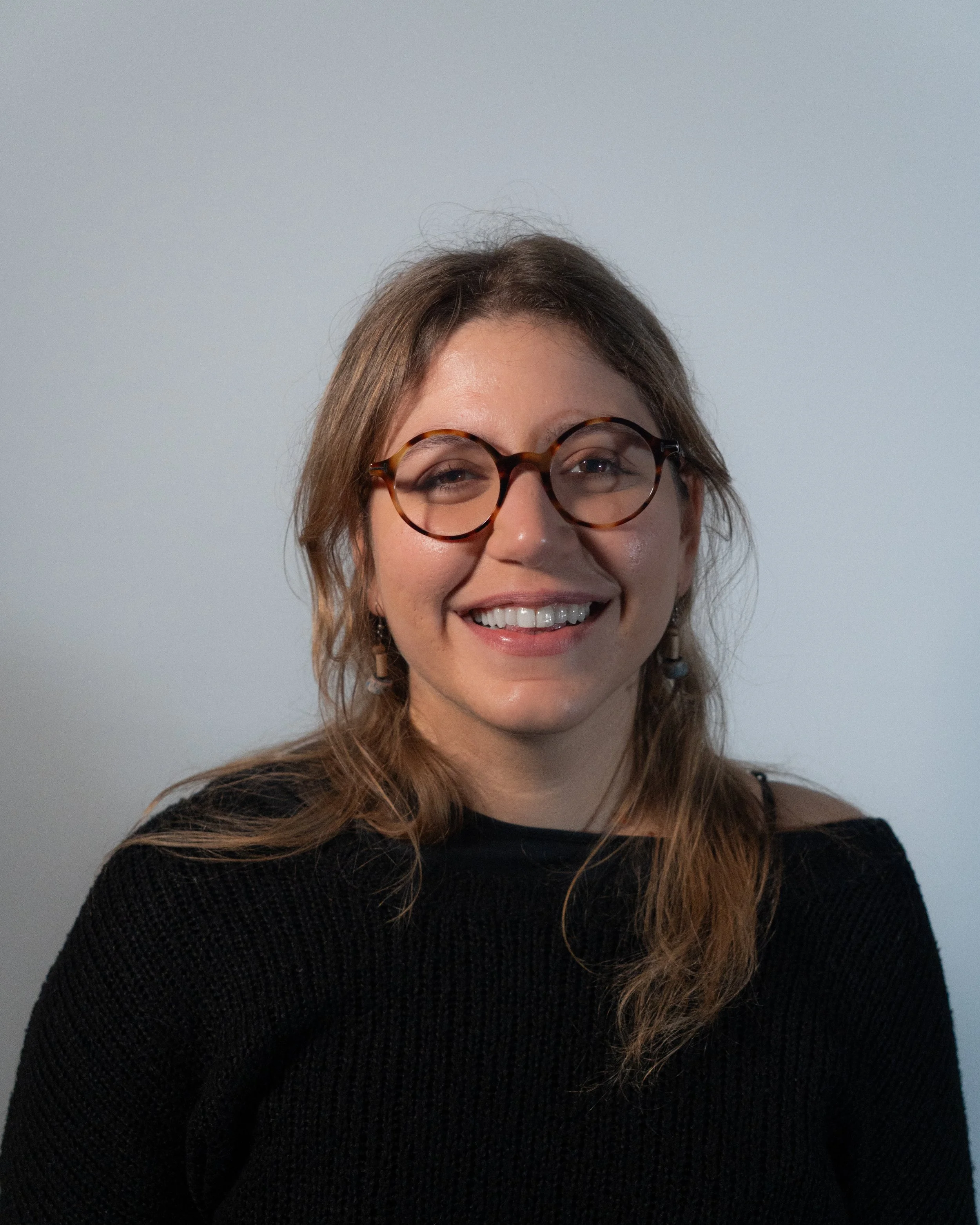 A woman with shoulder-length curly hair, glasses, and a striped shirt smiling in front of a plain wall.