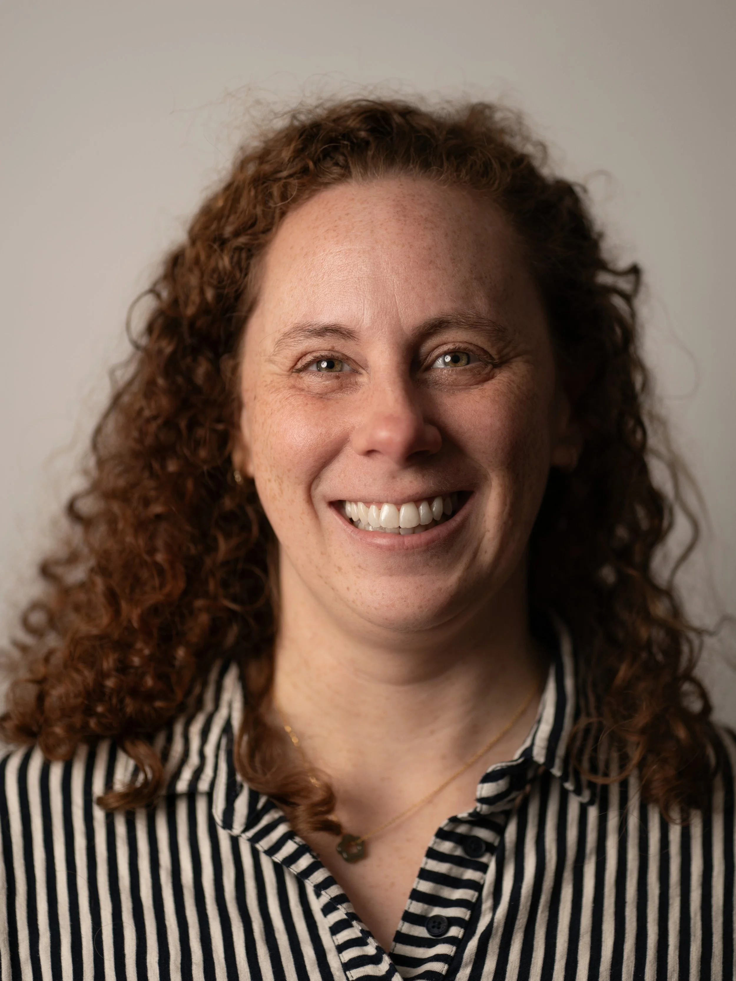 A young woman smiling with long, wavy hair, wearing a colorful sweater with pink and green patterns, standing against a textured neutral background.