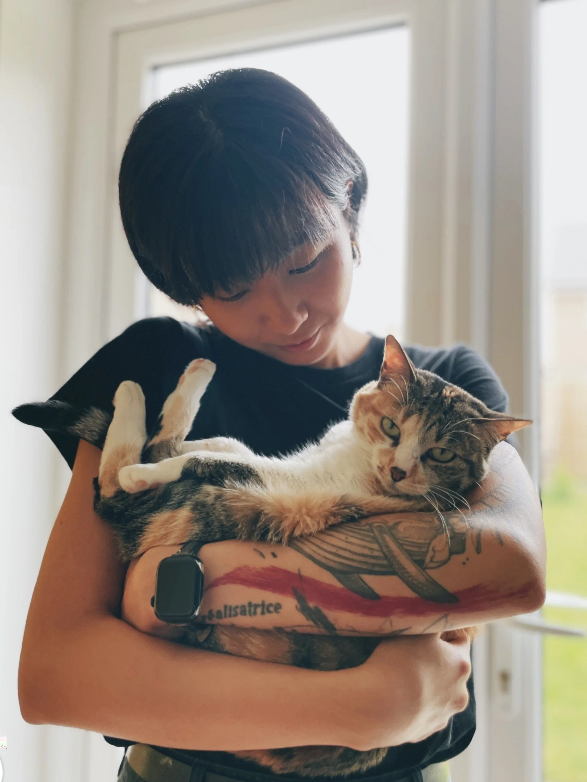 A person with short black hair holding a gray and white cat inside a room with a window in the background.
