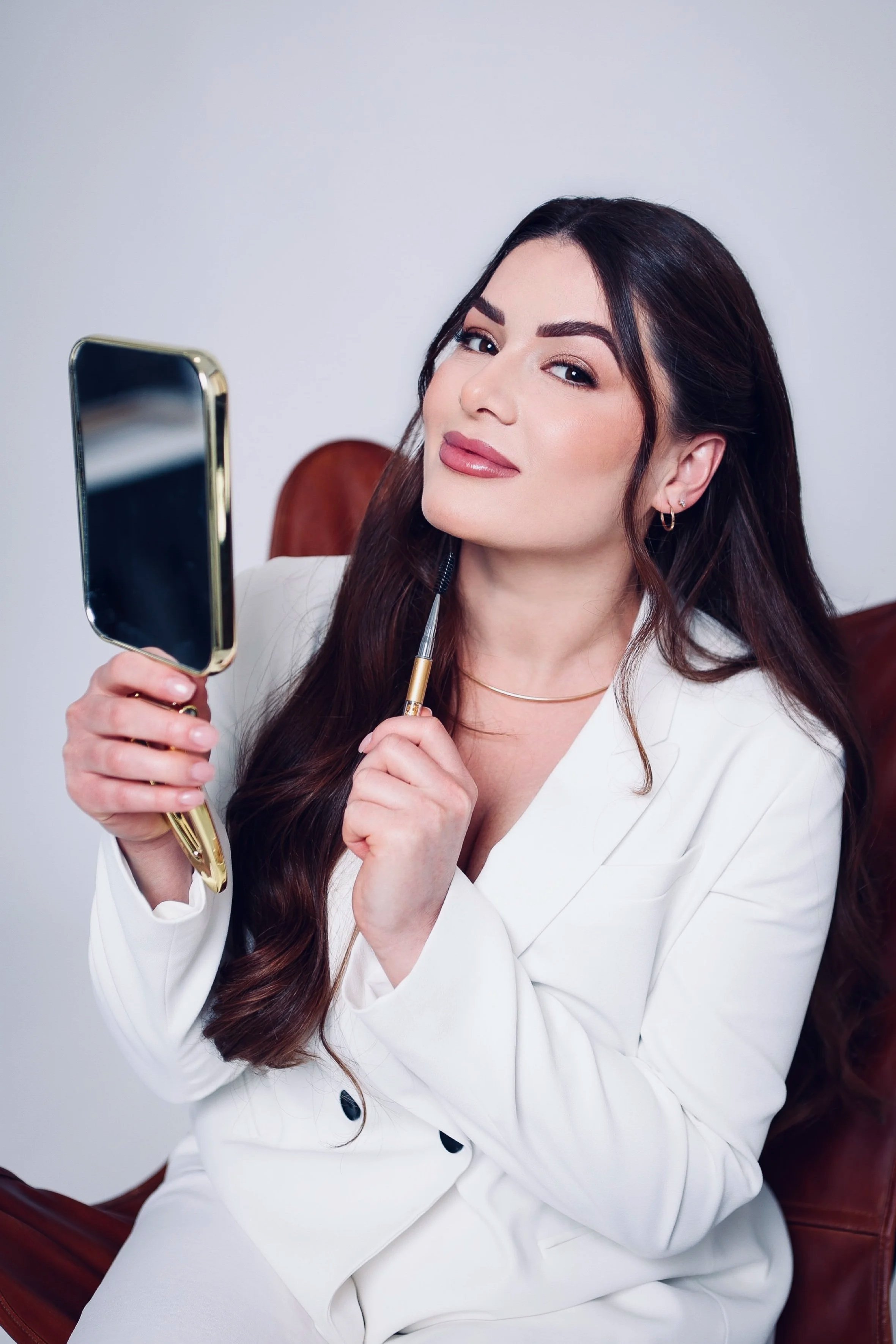A woman with long dark hair and makeup sitting on a leather chair, holding a mirror in one hand and a makeup brush in the other, posing for a selfie.