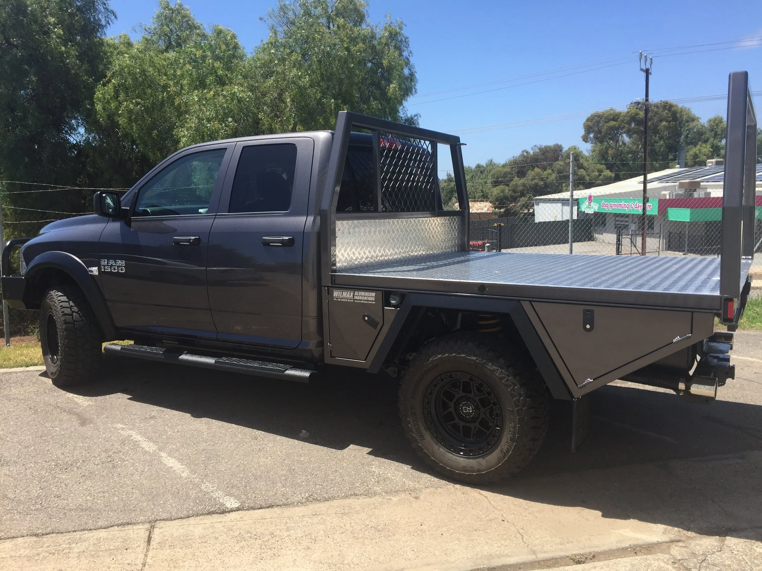 A dark gray flatbed truck parked on a street with trees and a building with a sign for dog grooming and daycare in the background.