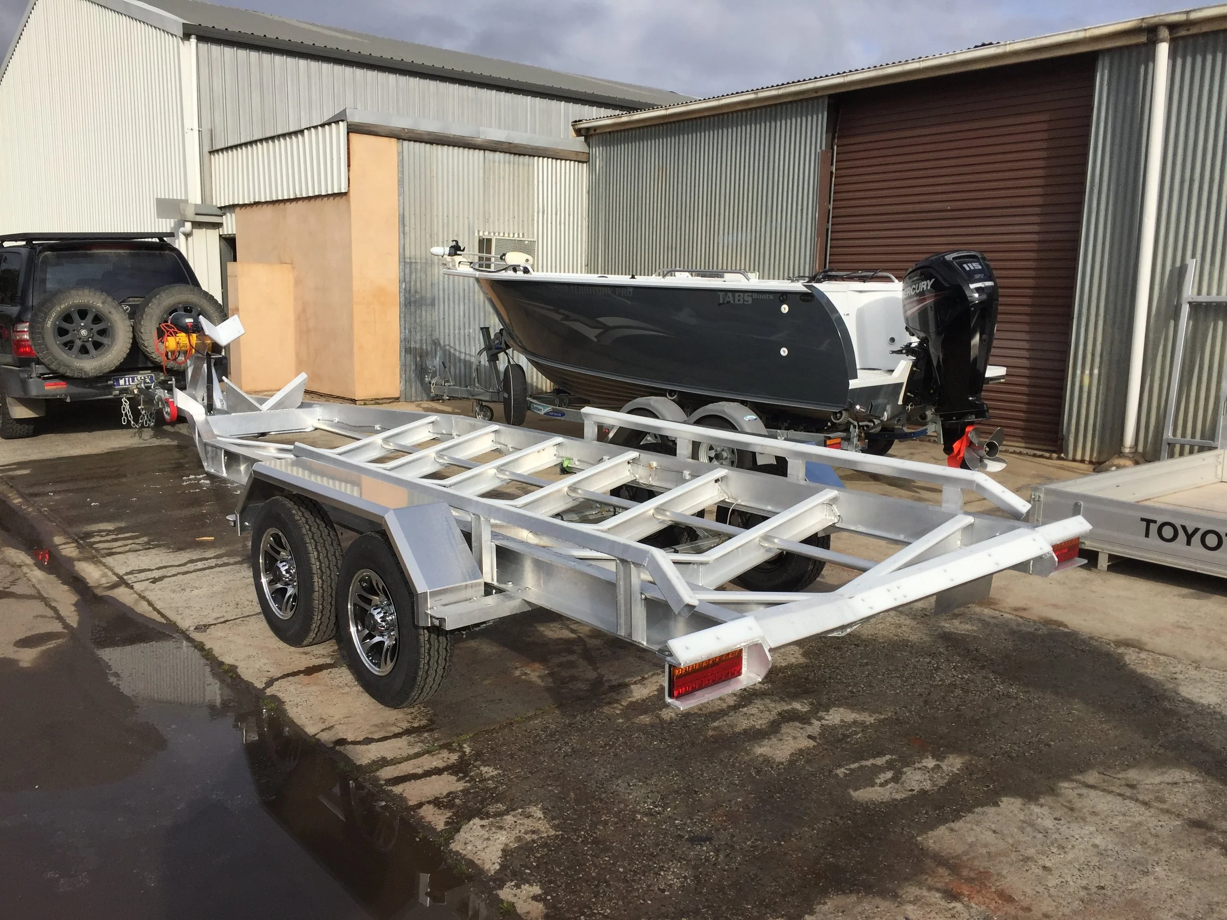 A boat on a trailer, parked outside a warehouse, with another trailer and two spare tires attached to a black vehicle nearby.