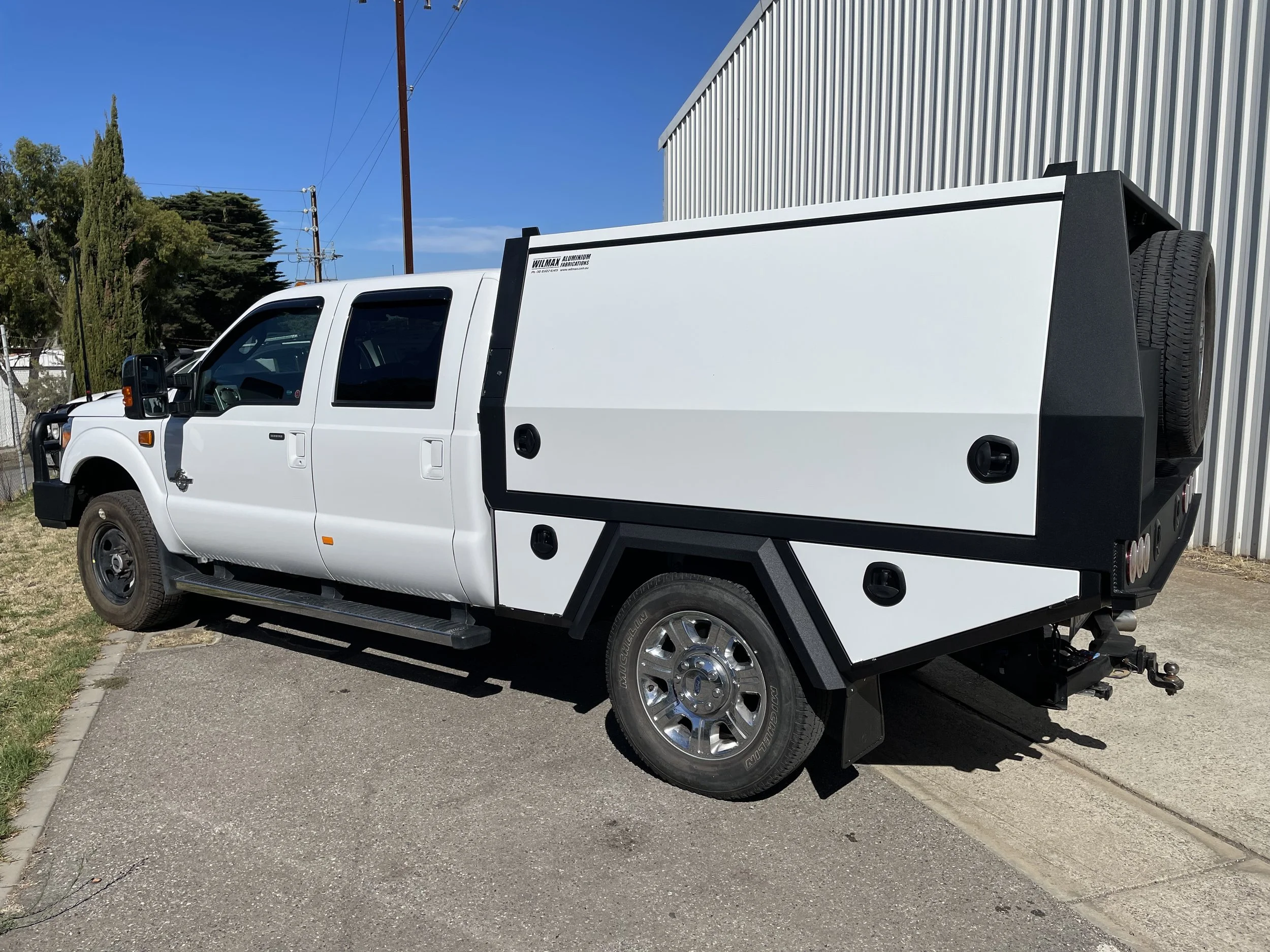 A white utility truck with a custom canopy parked beside a corrugated metal building, with trees and utility poles in the background and a clear blue sky.