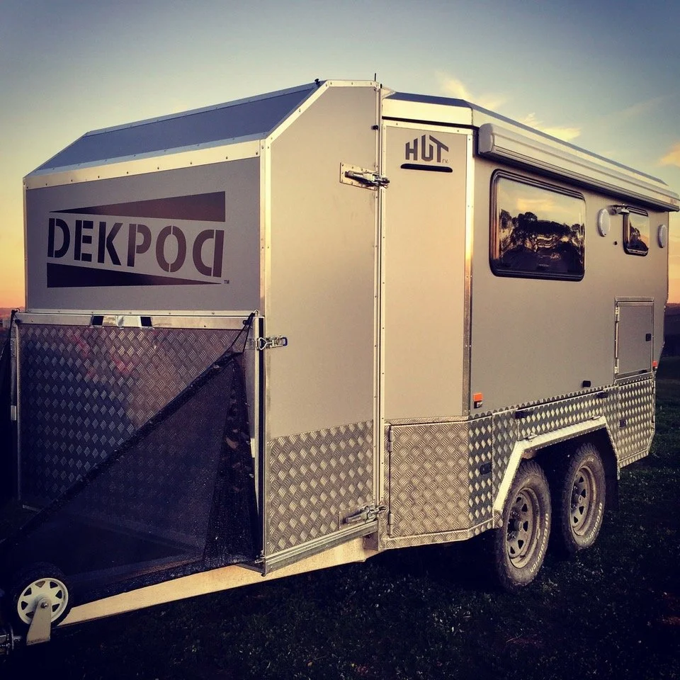 A silver travel trailer with black tires, windows, and a fold-out awning, parked on a grassy field during sunset.