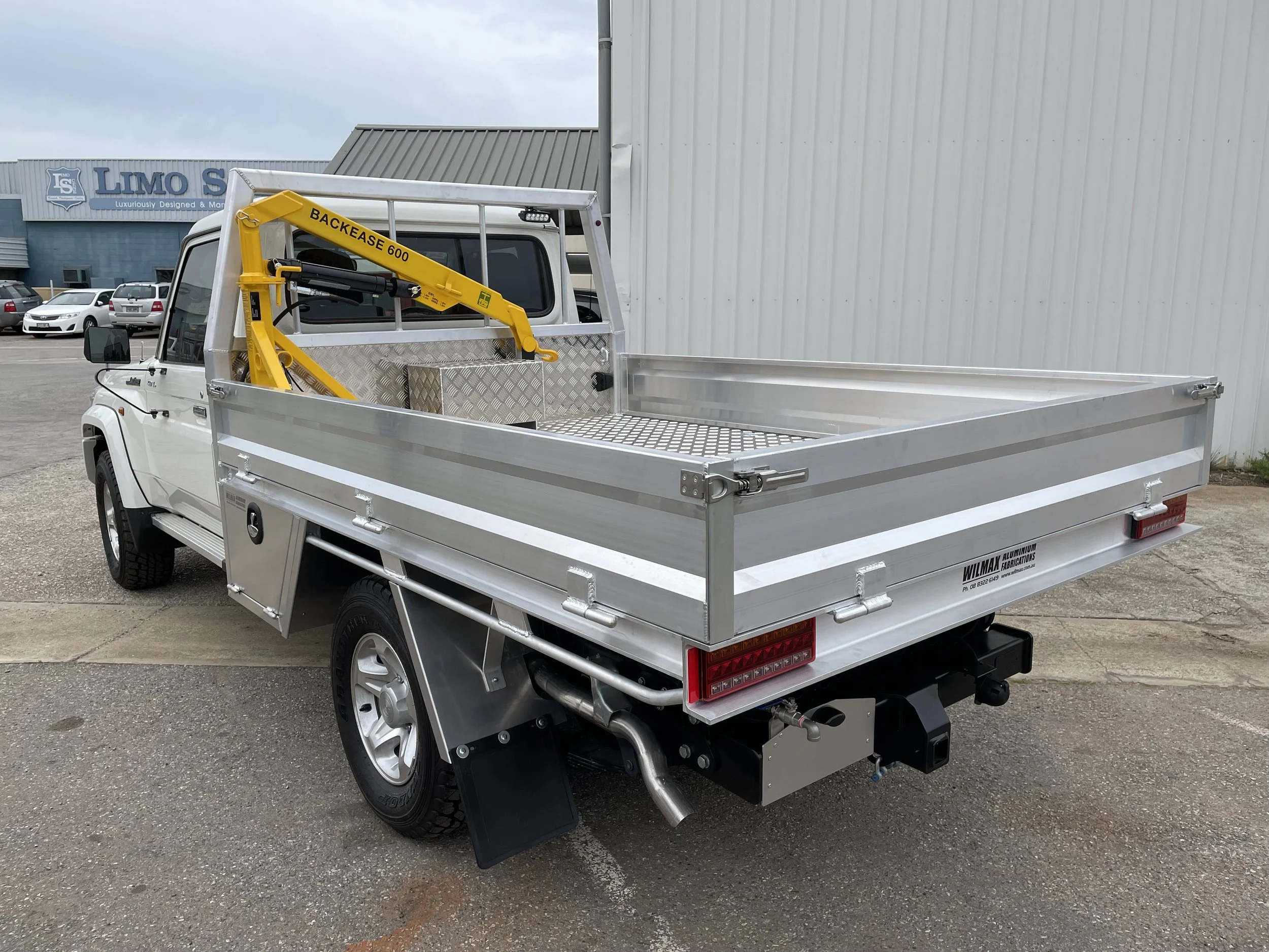 A white utility truck with an aluminum flat bed, The truck has a yellow backhoe loader attachment labeled "Backease 600" mounted behind the cab.