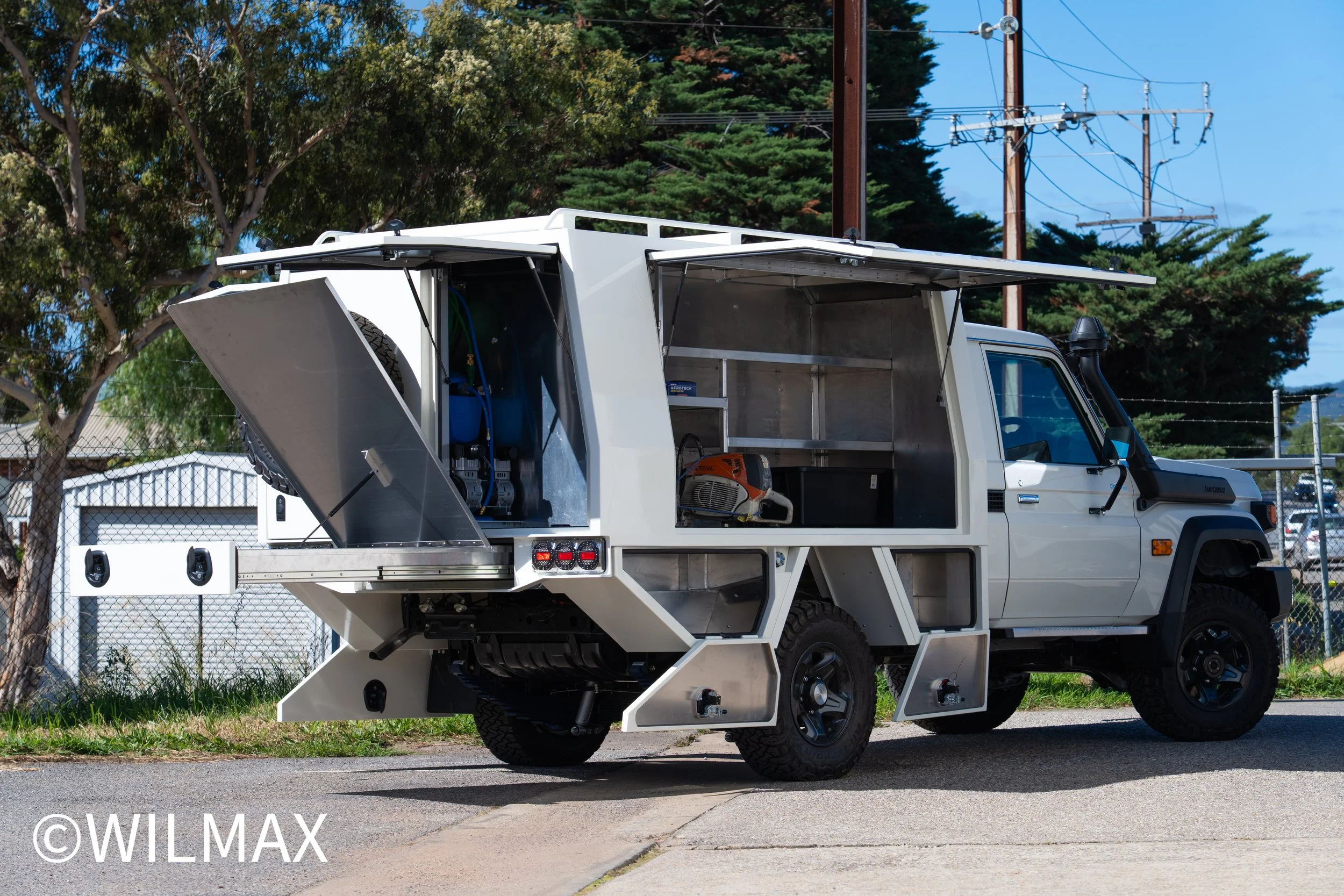 A utility truck with open storage compartments on the side, parked outdoors with trees and power lines in the background.