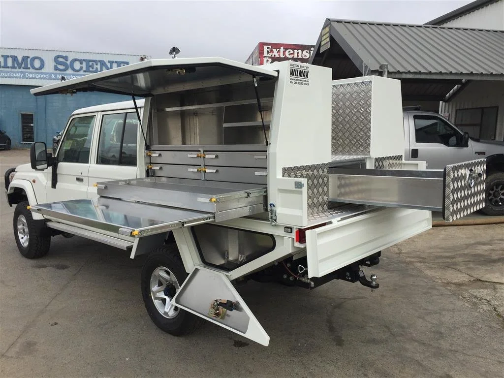 White utility truck with an open lift-up side storage compartment and additional compartments in the back, parked on a paved lot.