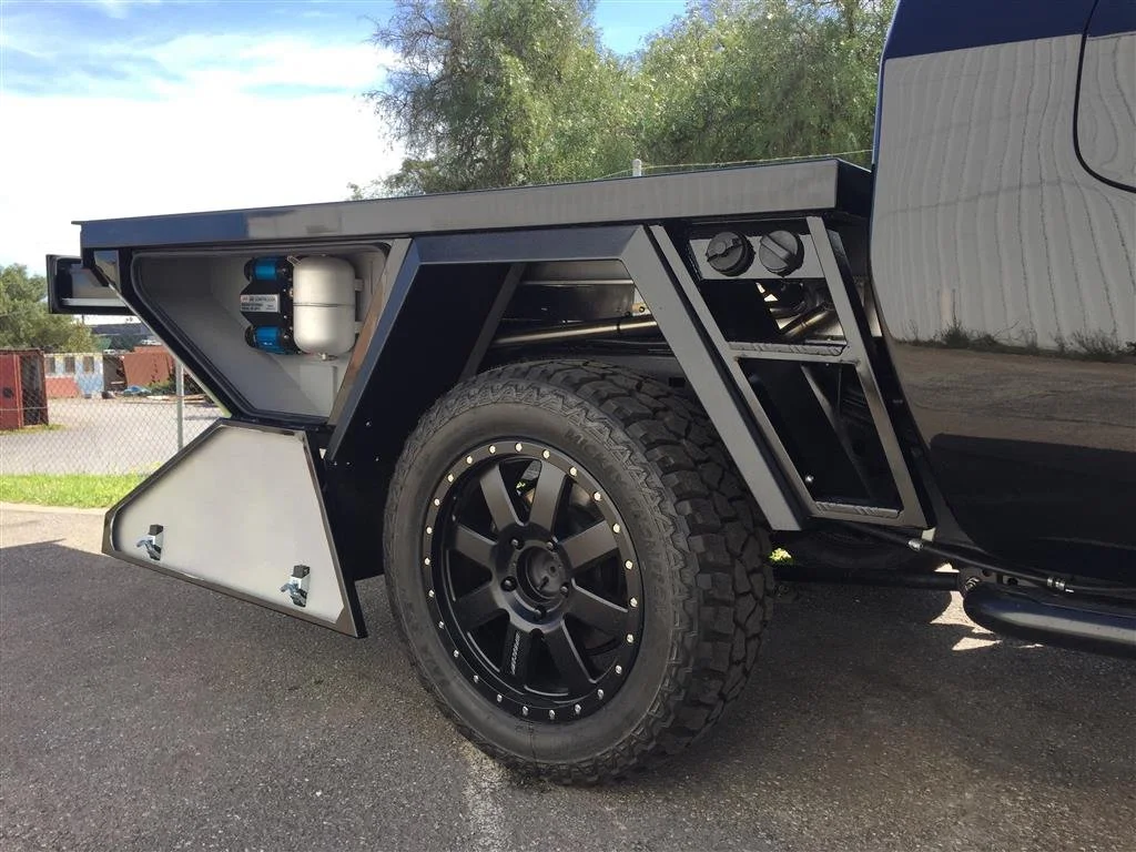 Close-up of the rear side of a black off-road vehicle with a specialized tire, black wheel, and an angular modern design. There is a storage compartment with a white panel and blue-capped filtration or compressor device inside, and multiple vents or openings on the body.
