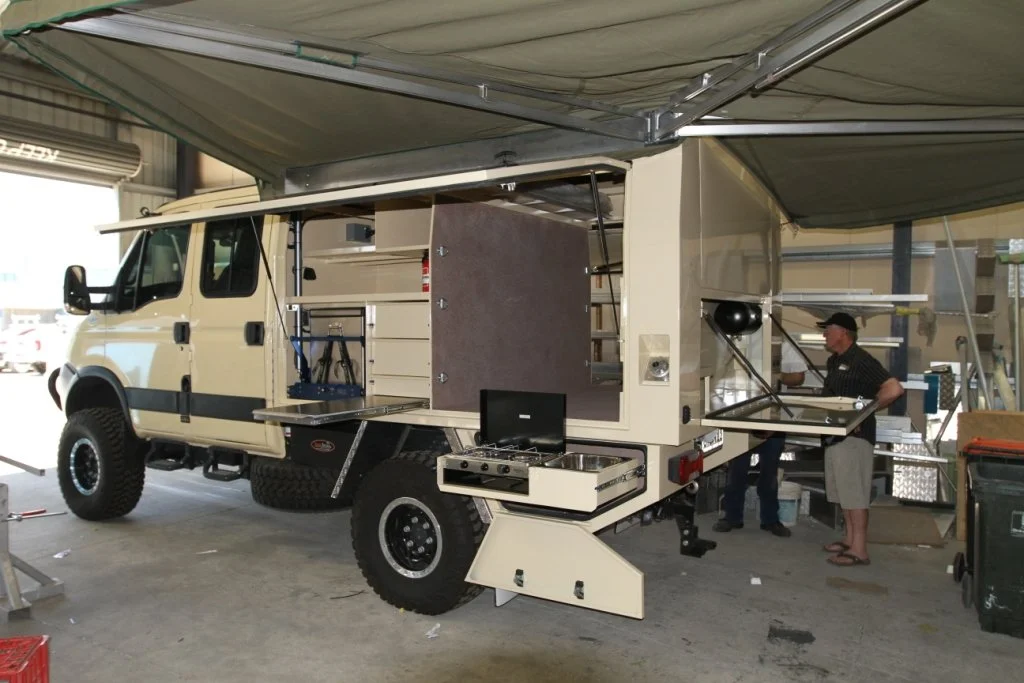 A custom-built mobile workspace or utility vehicle with a beige exterior, parked inside a garage with a partially open door. It has various equipment, storage compartments, and work surfaces, with two people working nearby.