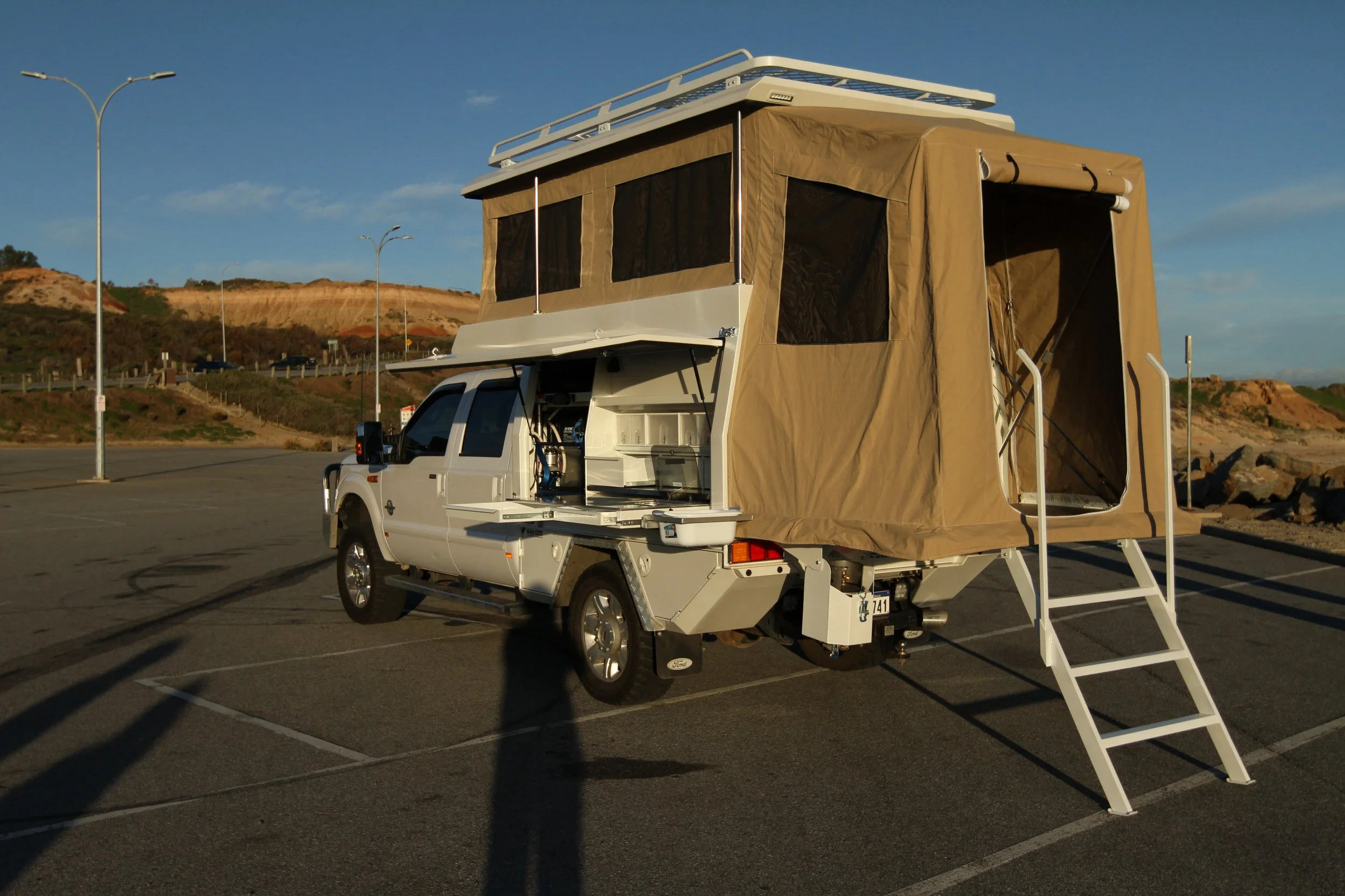 A white camper truck with a beige pop-up tent extension and stairs in an empty parking lot with streetlights and hills in the background.