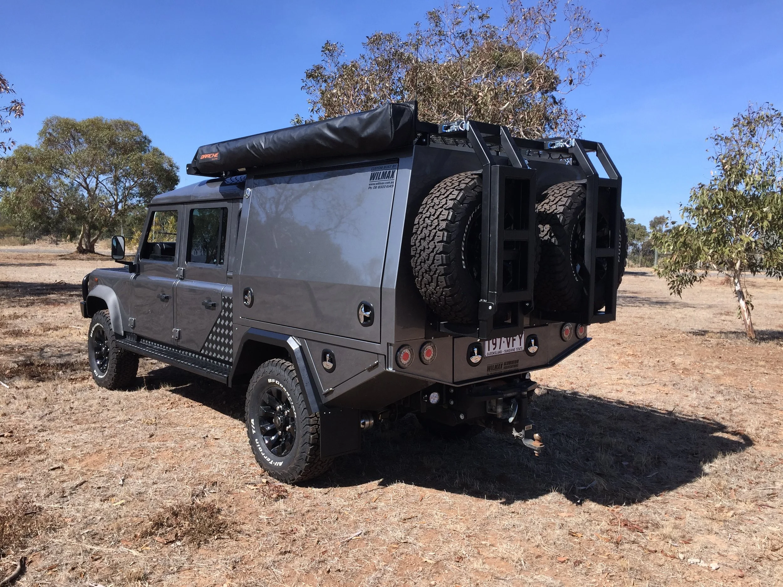A gray off-road vehicle with large tires custom canopy, a spare tire on the back, a roof rack, and a black cover on top, parked on dry ground with trees and a clear blue sky in the background.