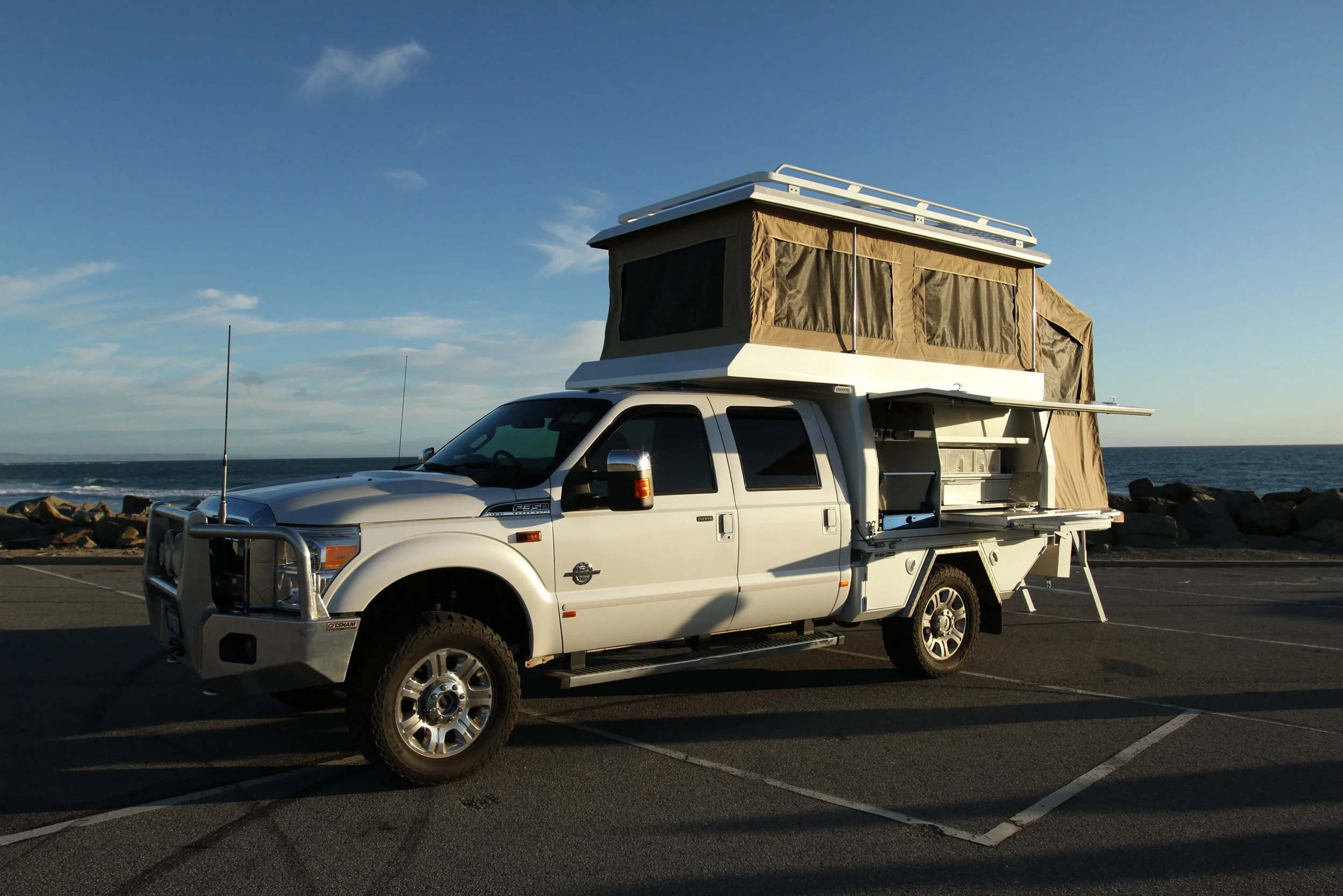 White pickup truck with a rooftop tent set up near the beach, parking lot, and ocean in the background under a clear blue sky.