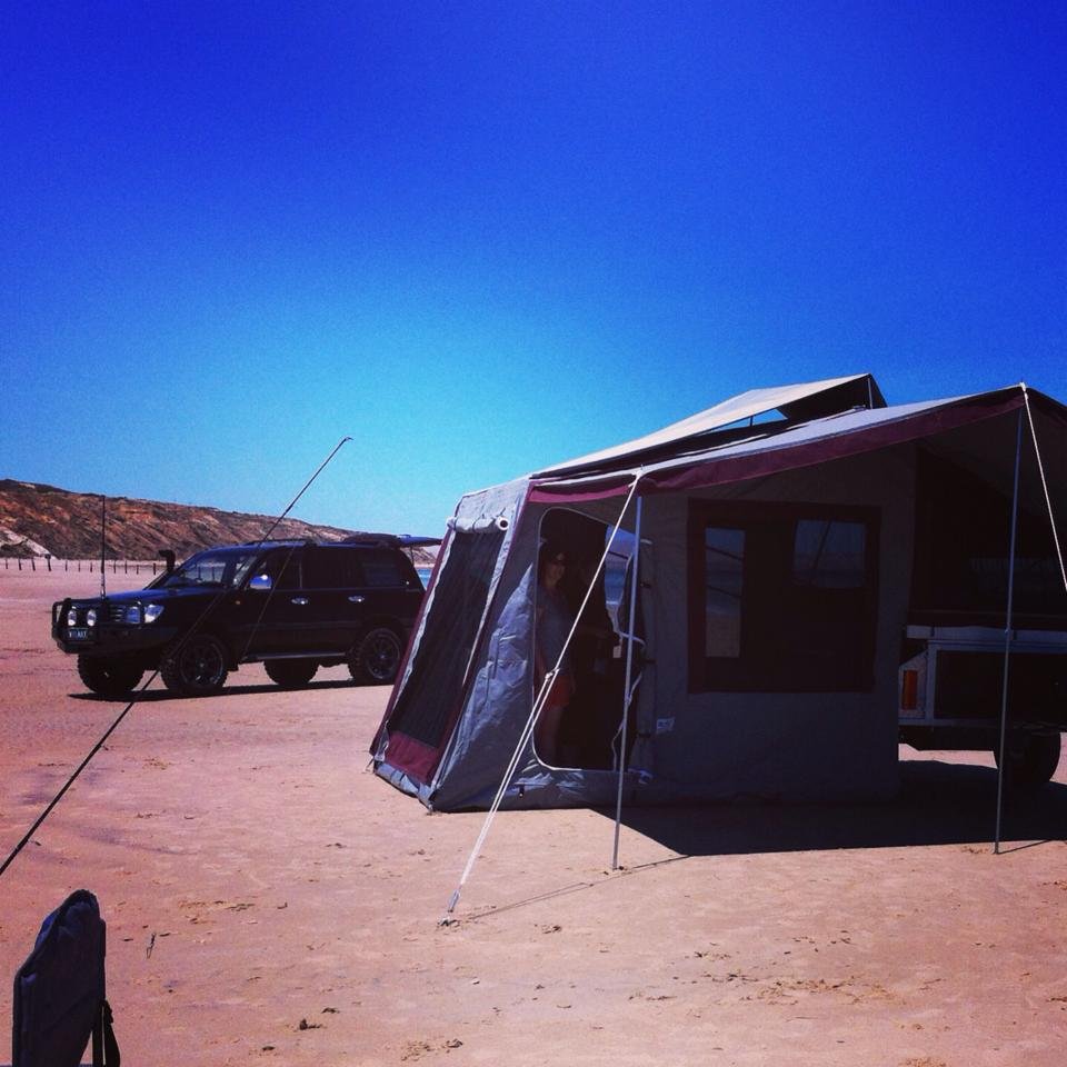 A black vehicle parked next to a camping trailer with an extended awning, set up on a dirt or sand terrain under a clear blue sky.