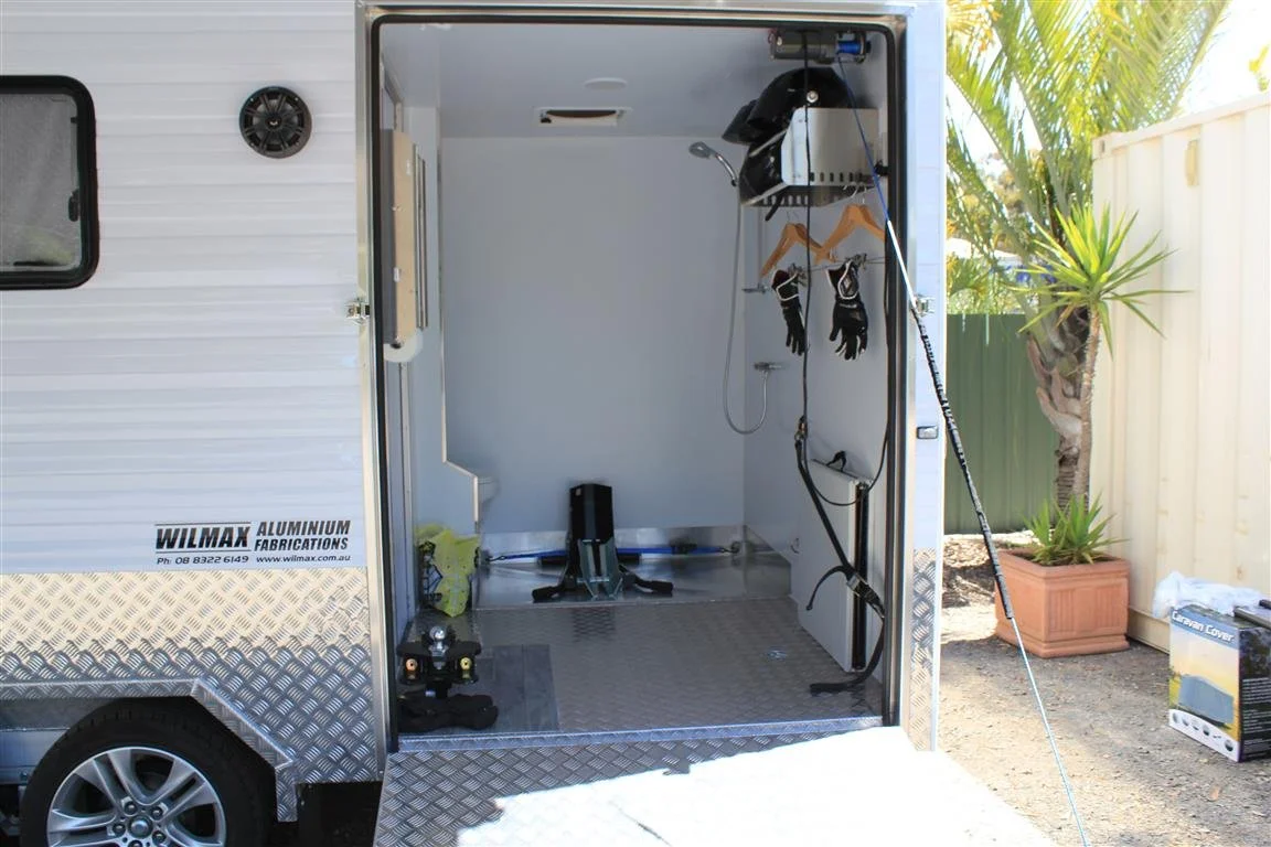 Empty garage inside a caravan with tools and equipment, hanging coat hangers, gloves, a step platform, and a showerhead on the wall, outdoors with a potted plant and fence nearby.