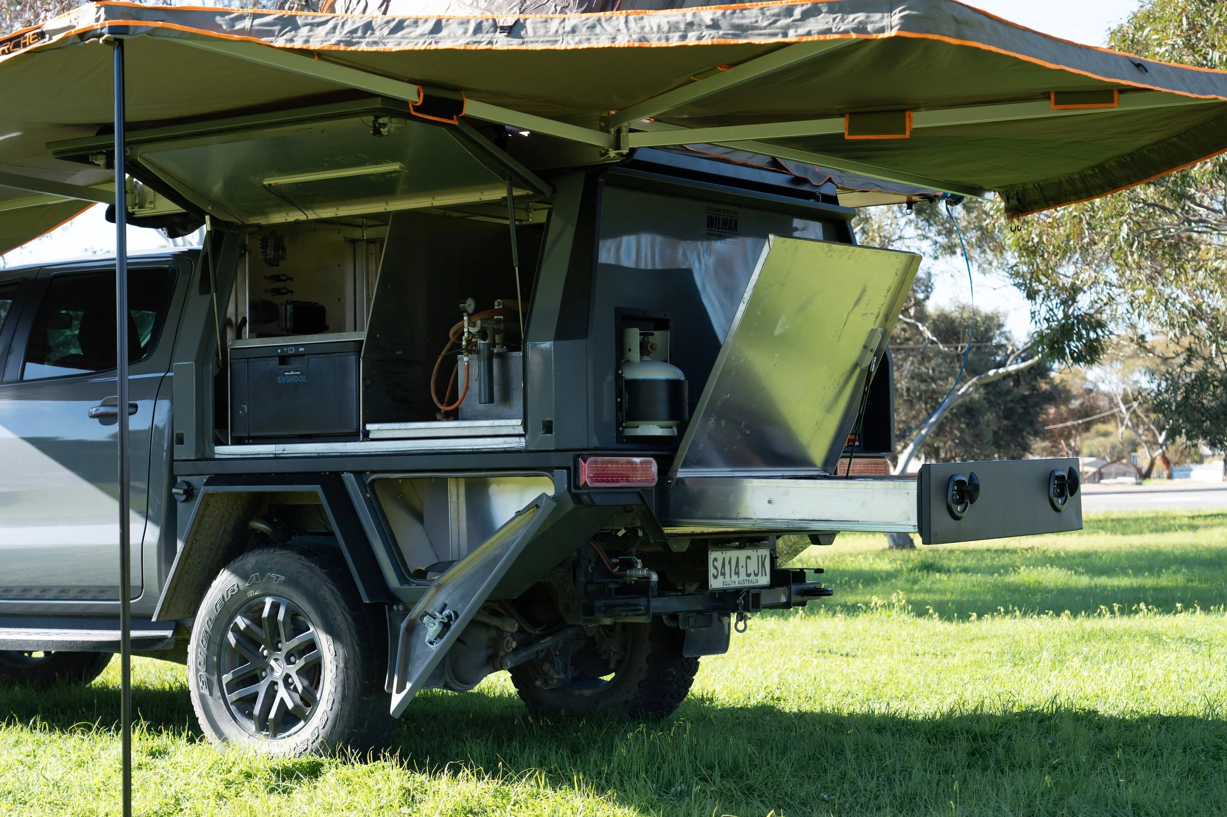 A custom canopy with a large awning extended in a grassy outdoor area, trees in the background, and a South Australia license plate.