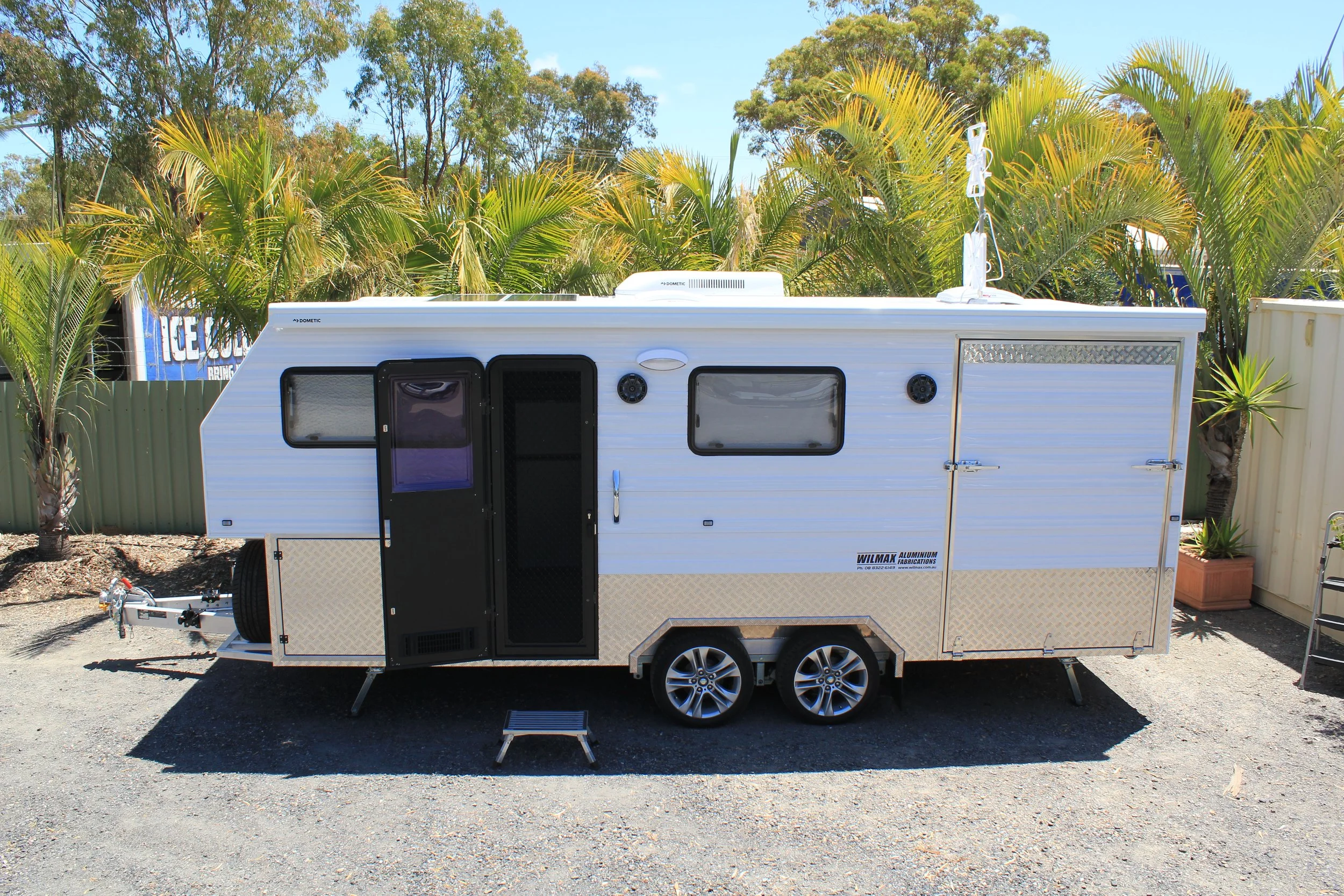 A white travel trailer parked on gravel with a small step ladder in front. There are palm trees and a blue sky in the background.