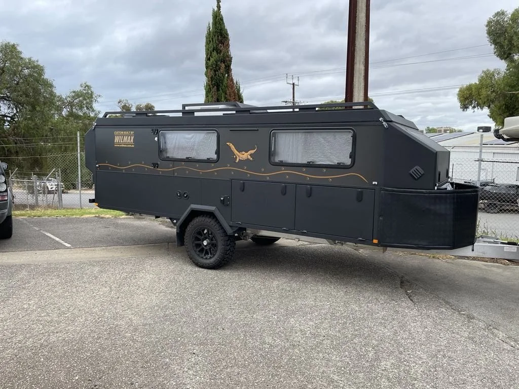 A black compact custom caravan with a lizard decal, parked in a lot with a chain-link fence and trees in the background.