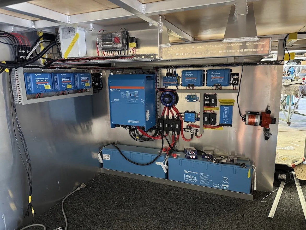 Electrical components and batteries inside a custom caravan, including blue lithium-ion batteries, circuit controllers, wiring, and a motor.