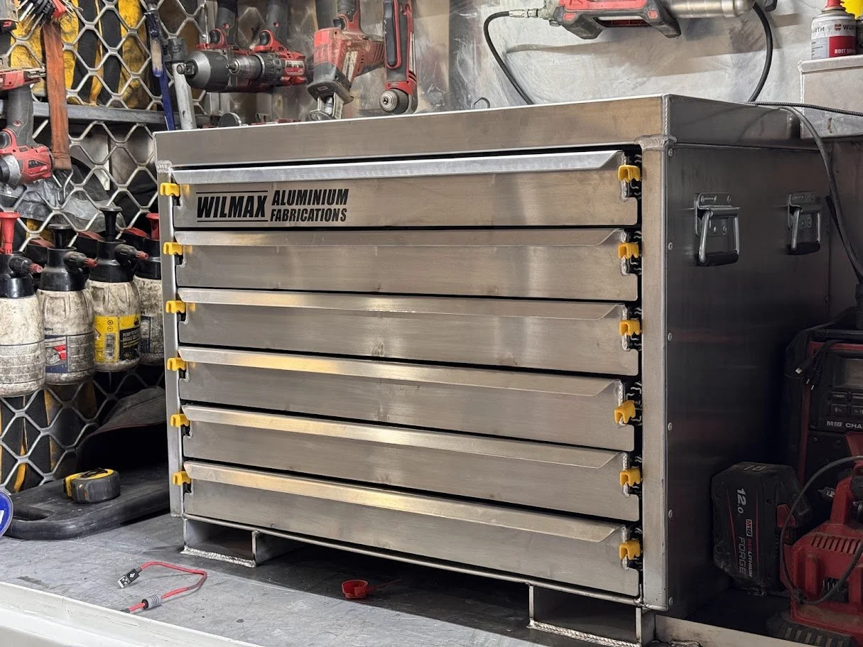 An aluminum tool storage cabinet with multiple drawers labeled 'WILMAX Aluminum Fabrications' in a workshop setting. The background shows various power tools, bottles, and workshop supplies mounted on the wall and placed around the workspace.