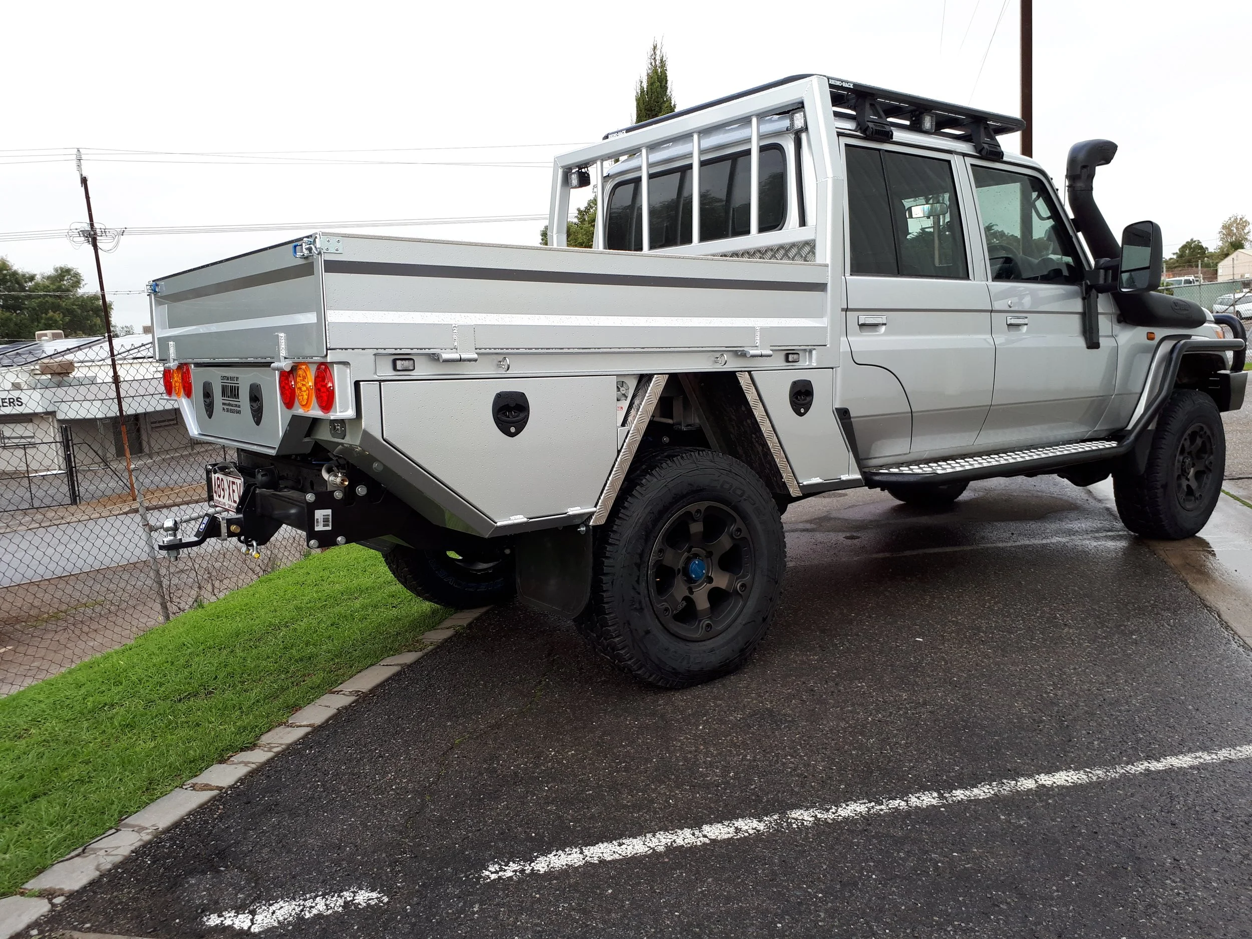 Silver 79 series landcruiser with a custom tray on a paved lot next to a grassy area, chain-link fence and utility poles visible in the background.