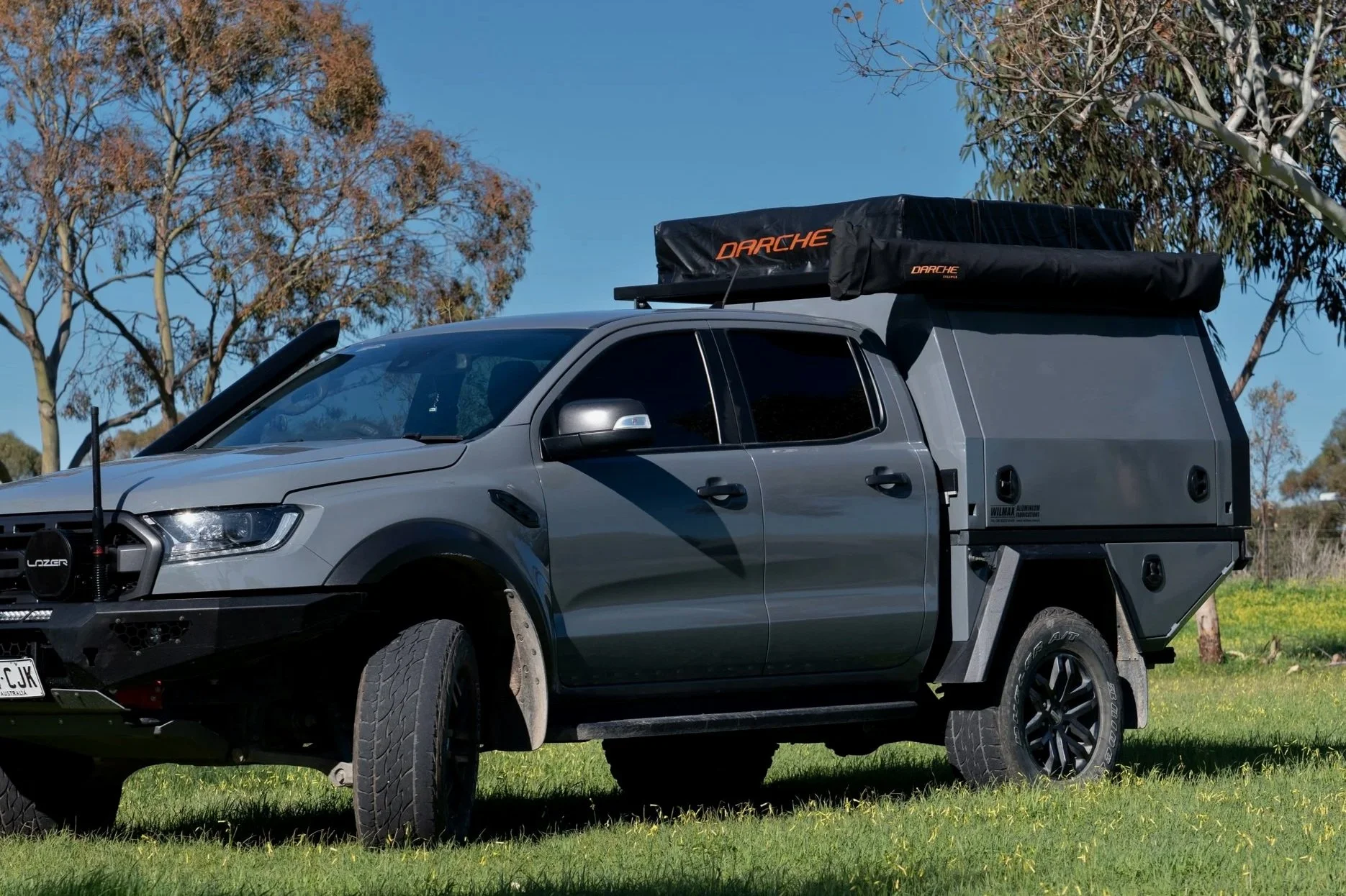 A gray ford ranger with a custom canopy camper attached to the bed, parked on grass with trees and a blue sky in the background.
