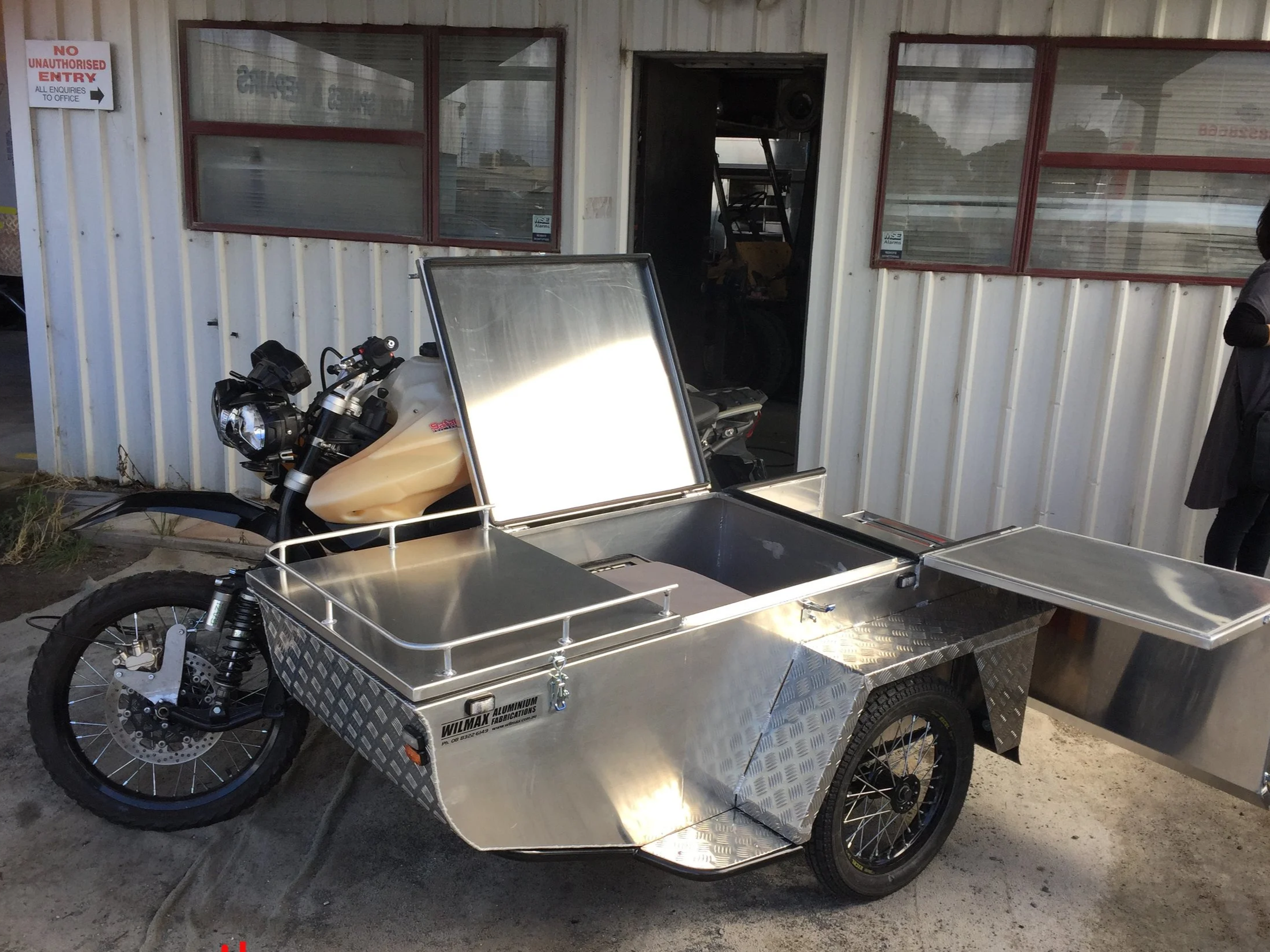 A motorized bike with a large aluminum storage box attached to the front, parked outside a building with a sign that reads 'No Unauthorised Entry.' The building has red-framed windows and a door with a speaker inside.