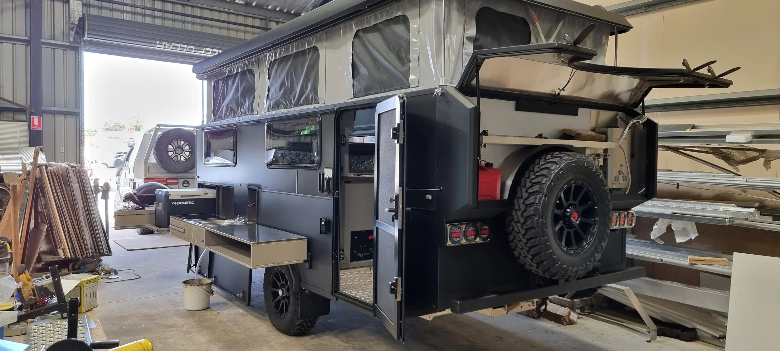 A black custom off-road caravan with a pop-up roof is inside a warehouse or garage, surrounded by various tools, equipment, and building materials.