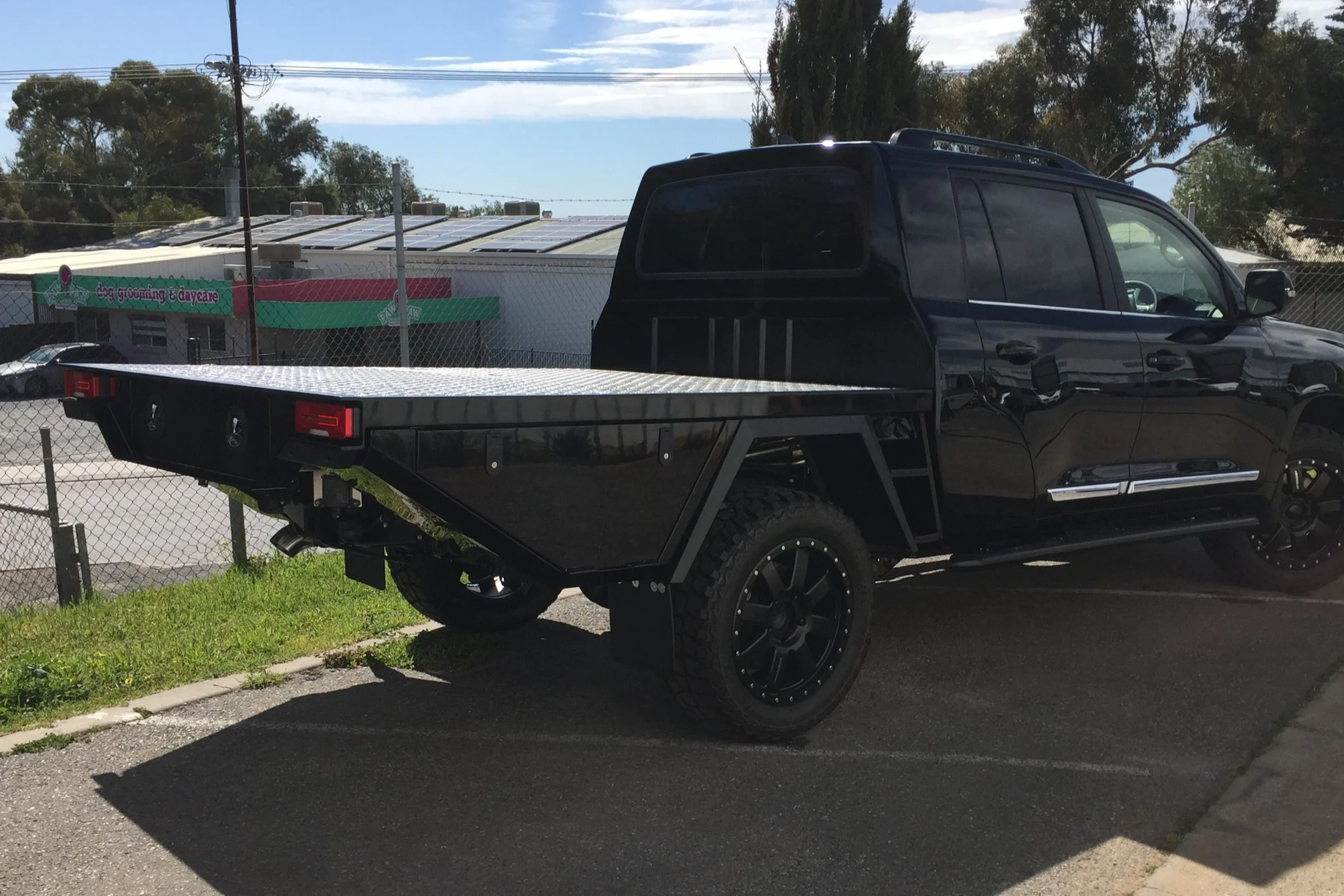 A black flatbed truck or ute tray parked on a street, with a building and trees in the background. custom tray on a landcruiser 200 series
