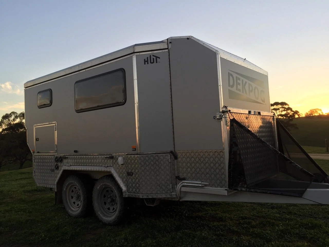 A silver custom camper trailer with black tires parked on grass during sunset, with trees and a hill in the background.