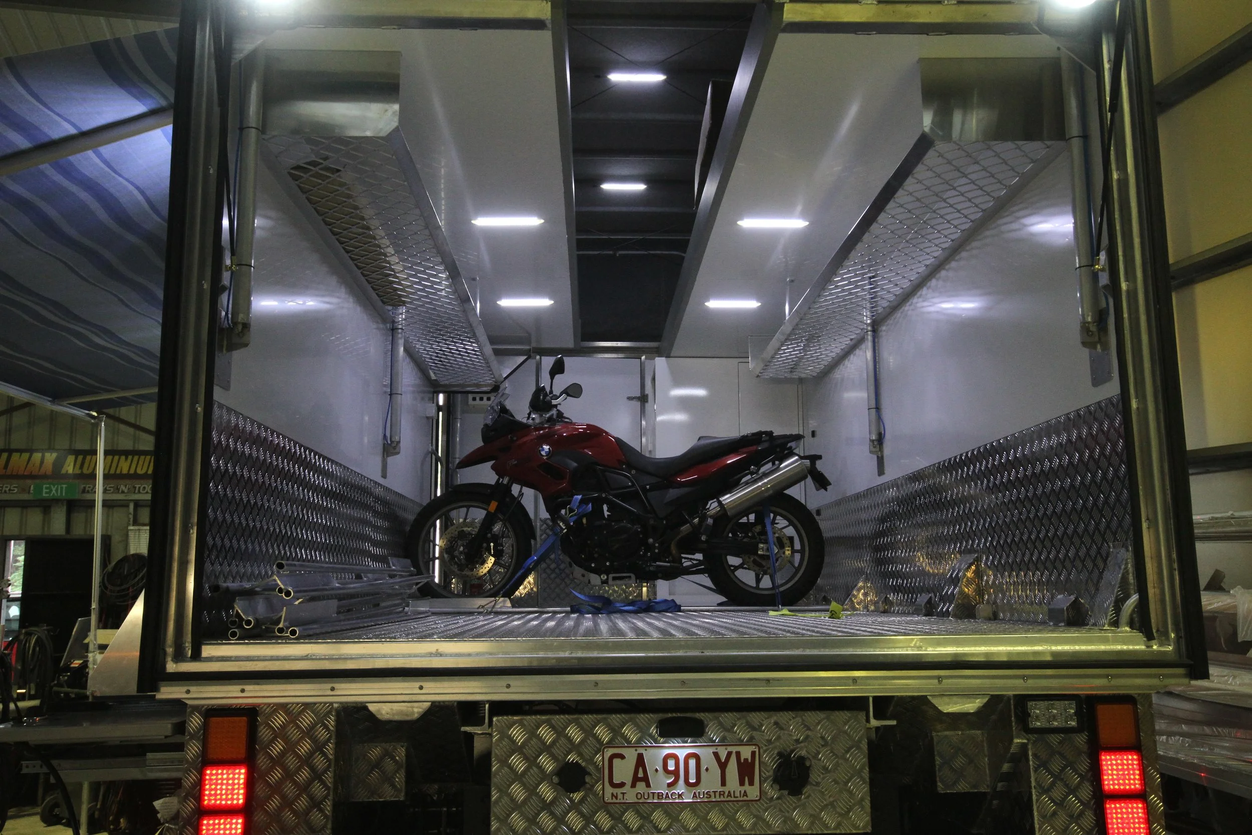 A red motorcycle inside a metallic enclosed trailer, with diamond plate panels and overhead lighting.