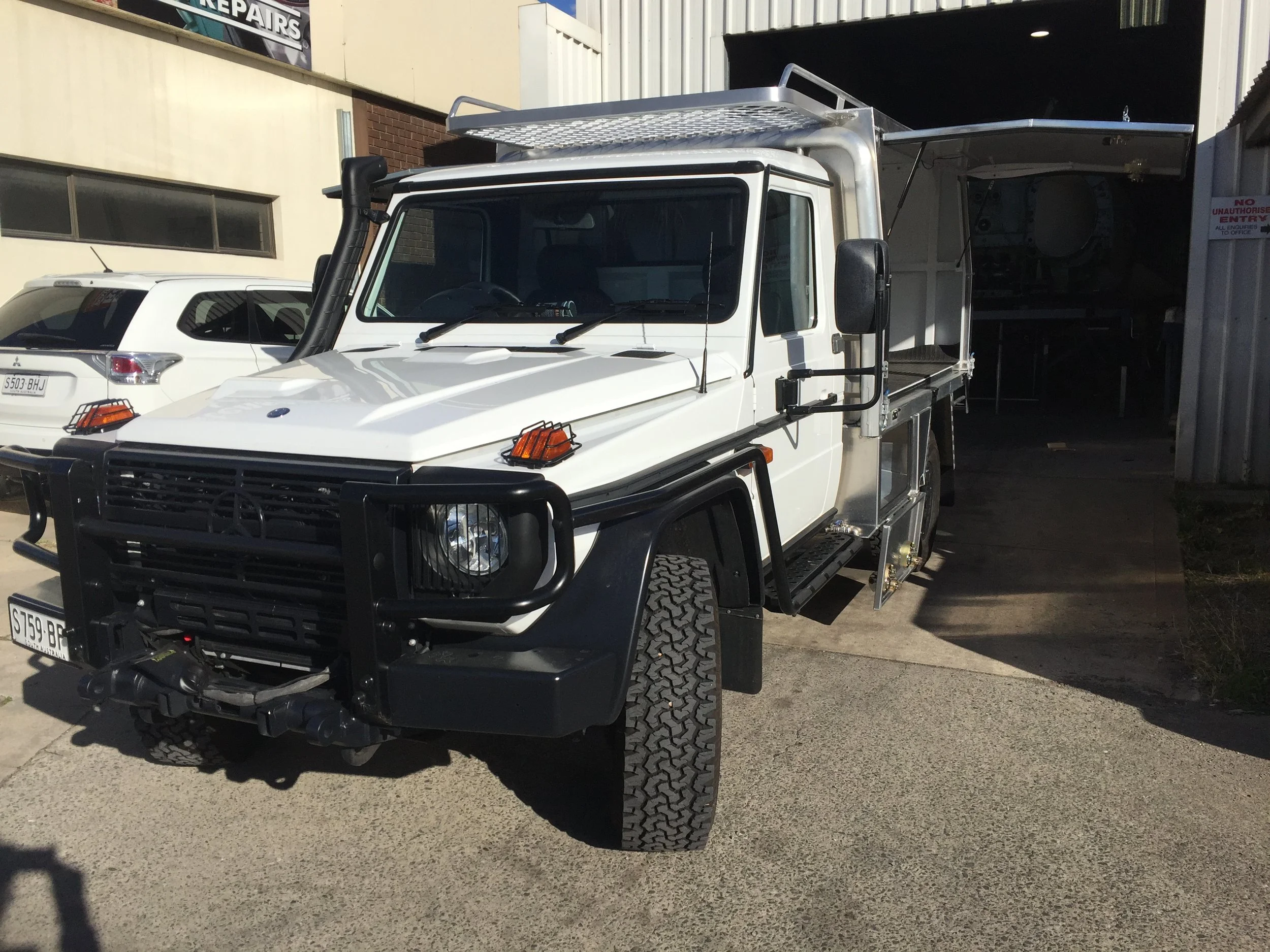 A white utility truck parked outside a building, with a large open cargo area and a ladder on the roof. There is a grey car behind it with a visible license plate.