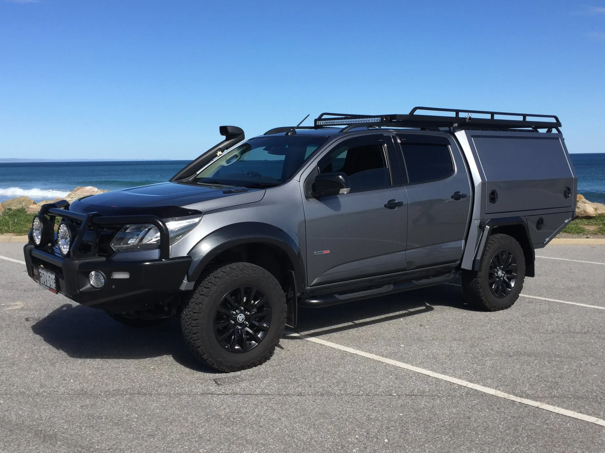 A silver pickup truck with off-road tires and accessories parked near the ocean, featuring a canopy, roof rack, and auxiliary lights on the front bumper.