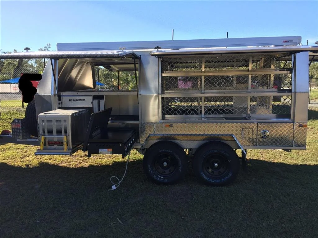 Metal food concession trailer with a mounted air conditioning unit, parked on grass near a sports field with a chain-link fence and trees in the background.