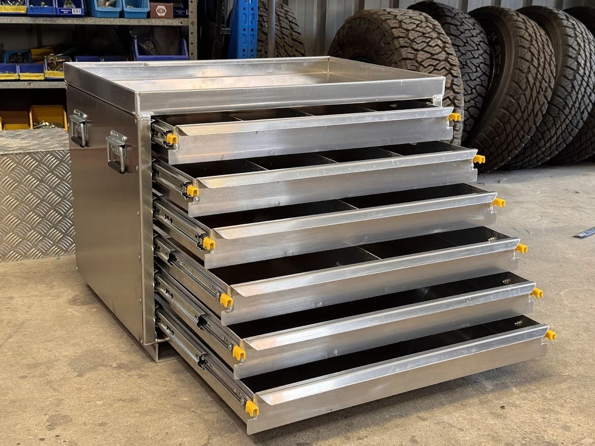 custom aluminium tool cabinet with multiple open drawers, in a garage or workshop setting, with vehicle tires stacked in the background.