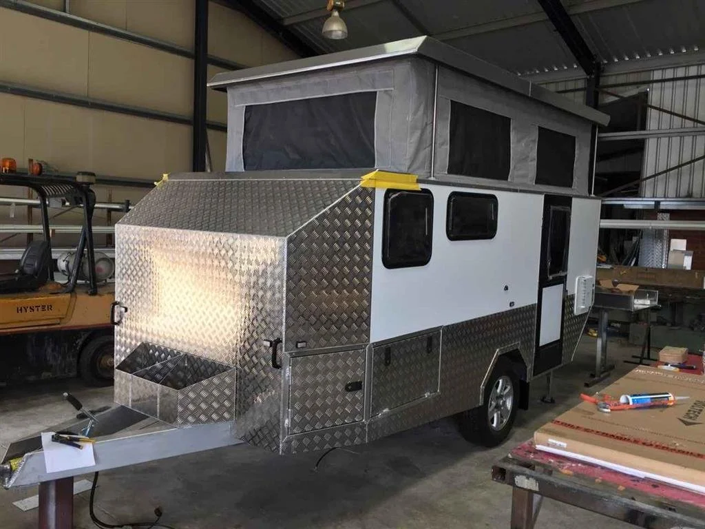 A small custom camper trailer with a pop-up roof inside a warehouse, with metal diamond plate on the front and lower sides, black window screens, and a door on the side.
