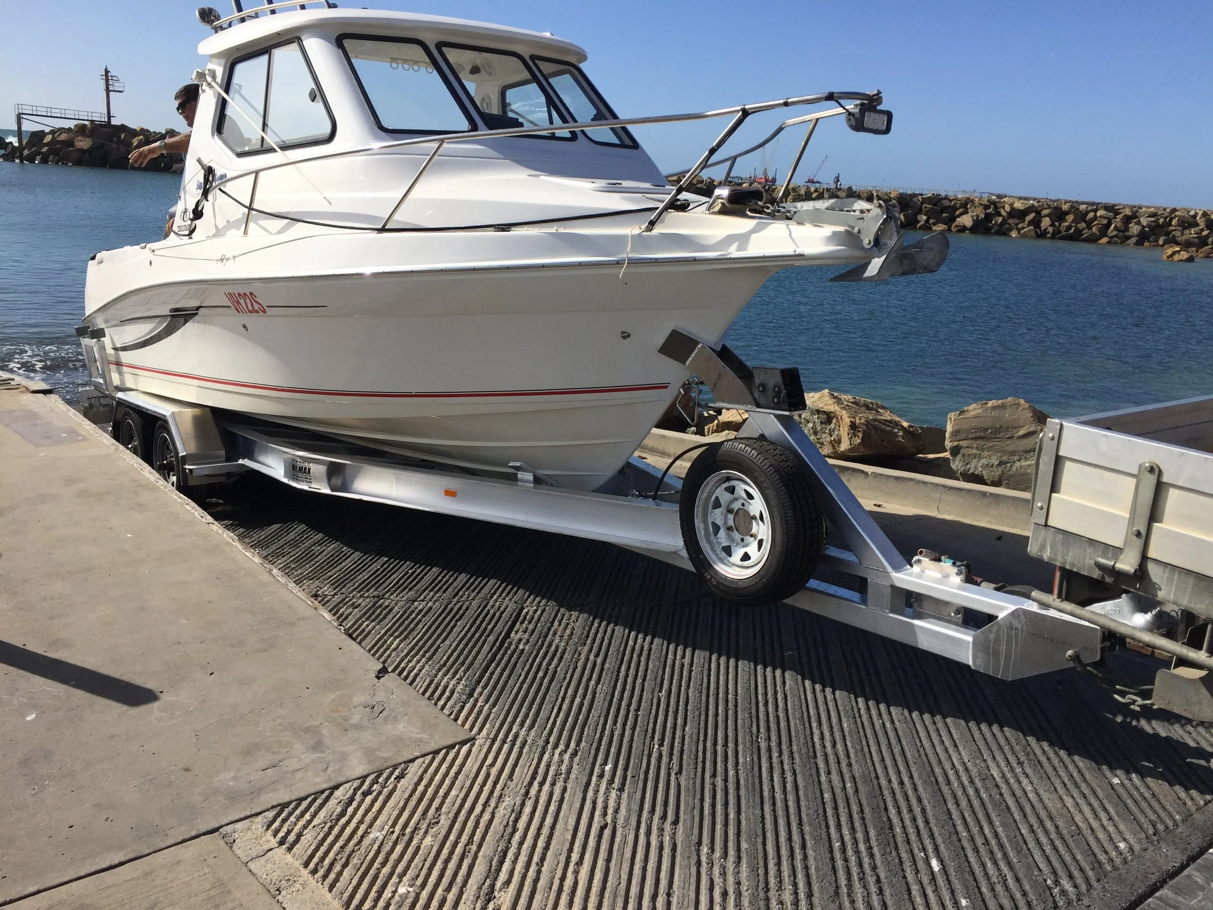 A white motorboat is on a trailer at a dock, with a man inside looking out. The background features a rocky breakwater and a calm body of water under a clear blue sky.